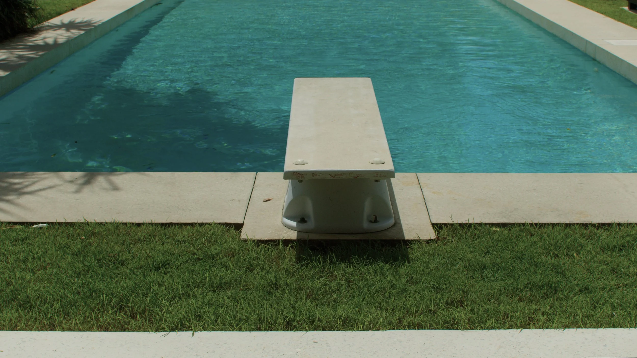 Empty diving board at a swimming pool with green grass and white concrete surrounding it.