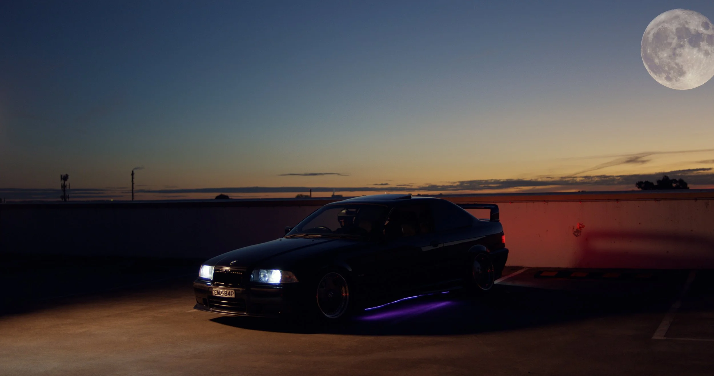 Black sports car with purple underglow lights parked on a rooftop parking lot during dusk, with a full moon and a clear sky in the background.