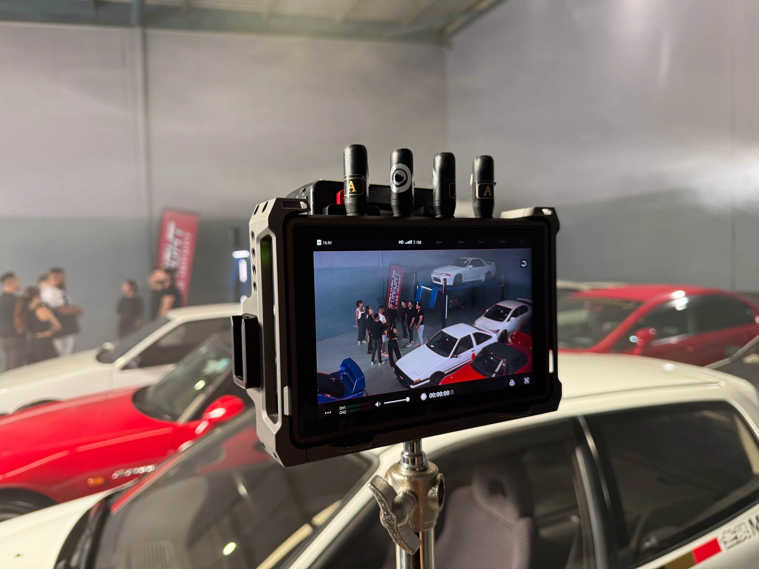 Camera monitor capturing a scene of an indoor car exhibition with a white sports car and a group of people gathered around it.