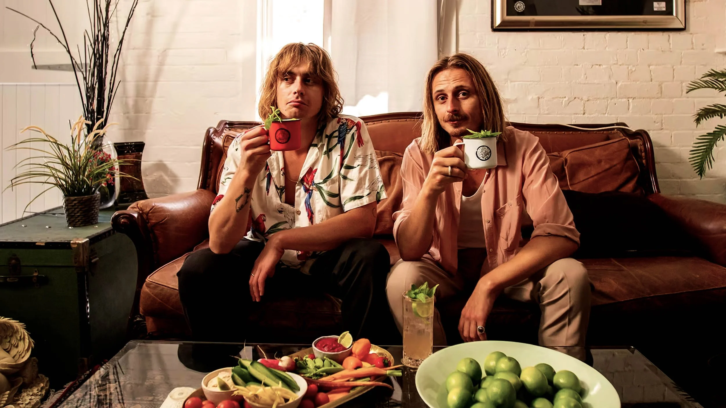 Two men sitting on a vintage-style sofa, holding mugs and surrounded by fresh fruits and vegetables on a coffee table, in a cozy, well-lit room with plants and white brick walls.