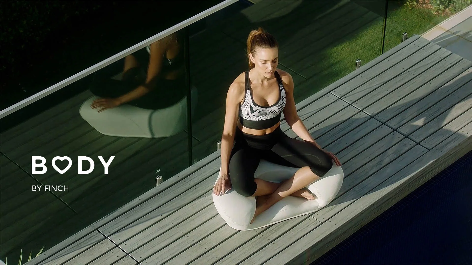 A woman practicing yoga outdoors on a wooden deck, sitting cross-legged on a cushion with her eyes closed, wearing athletic wear. There is a glass wall behind her, reflecting her image.