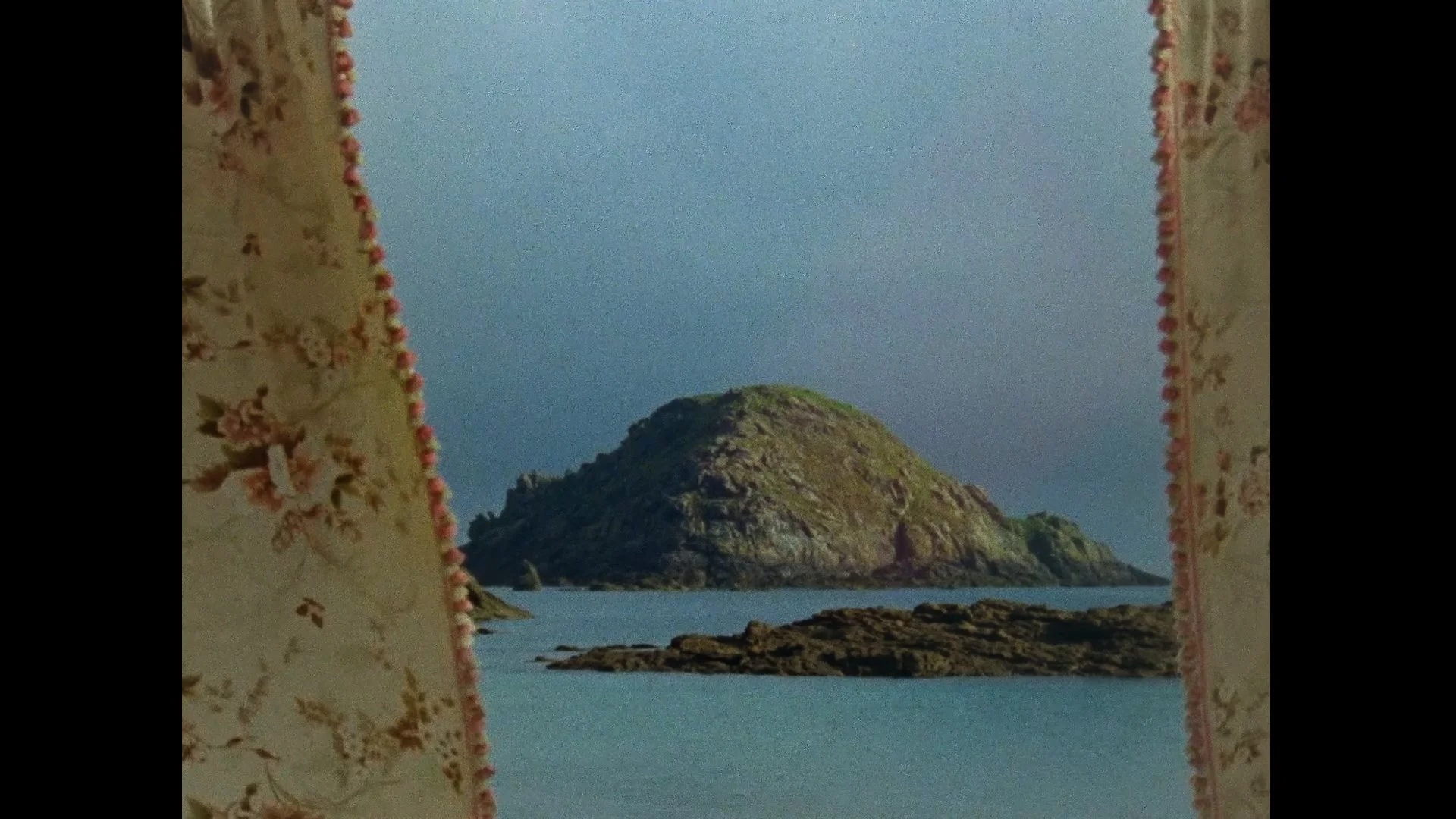 View of a rocky island surrounded by water, seen through a window with floral curtains.
