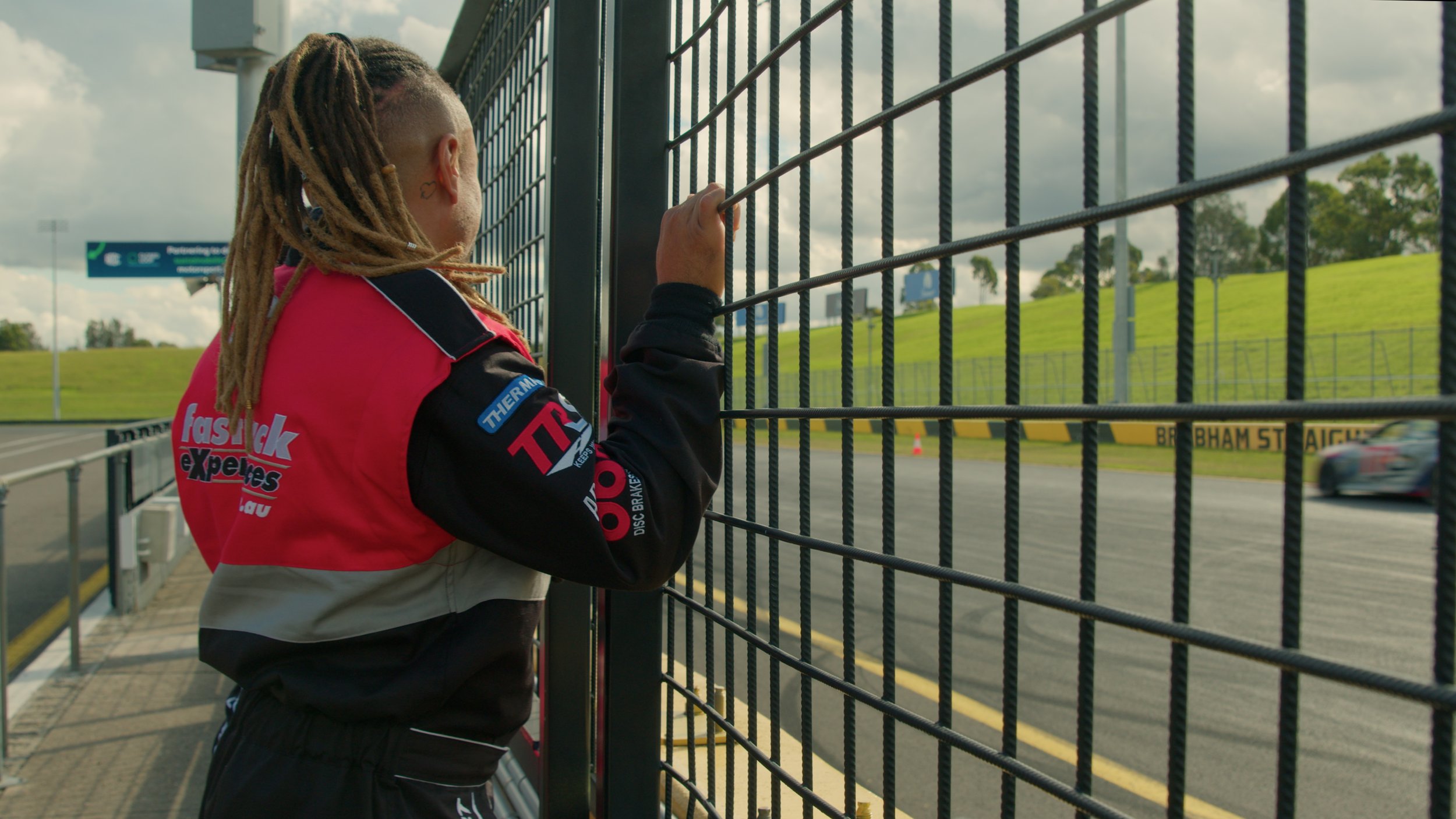 A person with dreadlocks wearing a racing suit and a red vest standing behind a metal fence, looking at a race car on a racetrack.