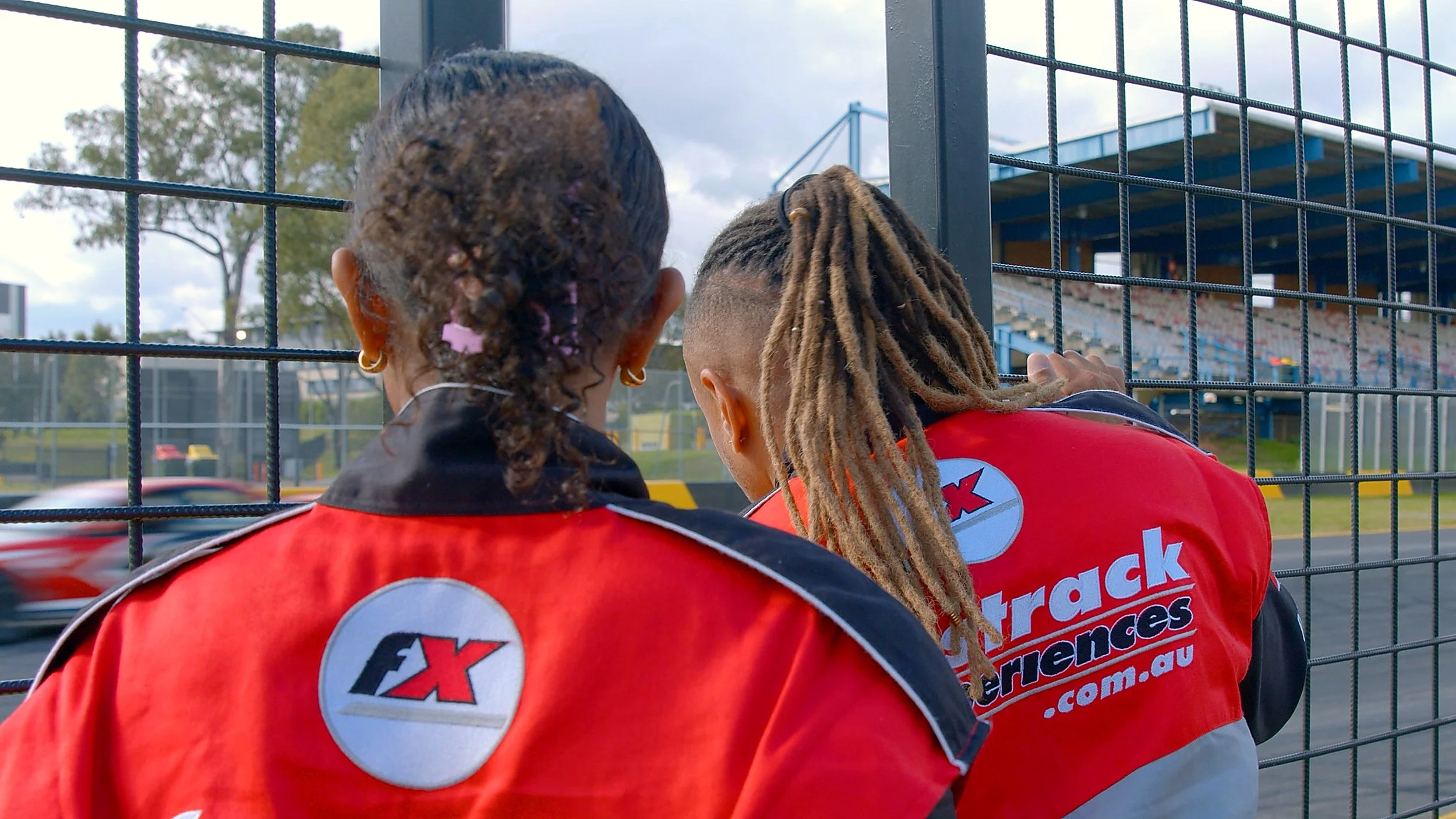Two individuals in racing uniforms leaning against a fence at a race track, watching a car pass by.