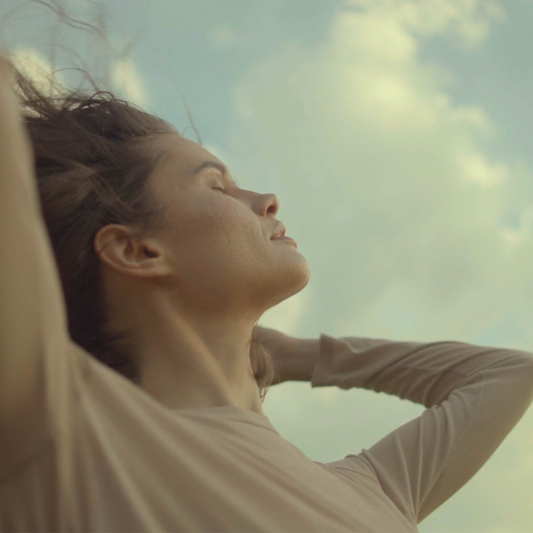 A woman with closed eyes relaxing outdoors with a blue sky and clouds in the background.