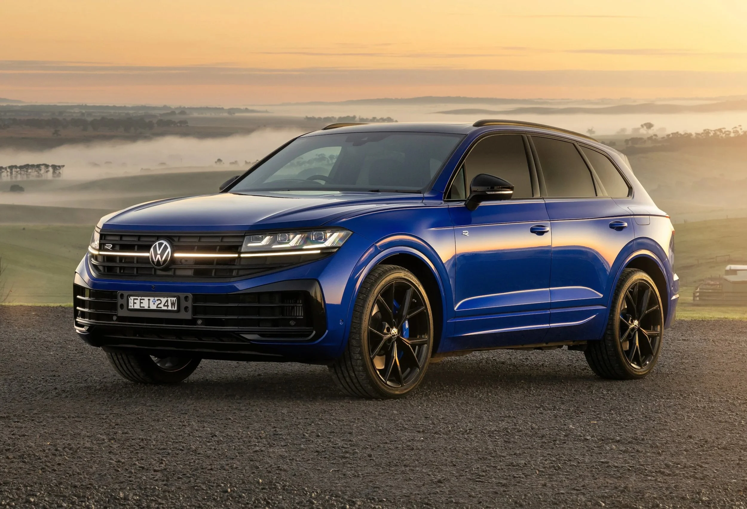 A blue Volkswagen SUV parked on a gravel surface with a landscape of rolling hills and fog in the background during sunrise or sunset.