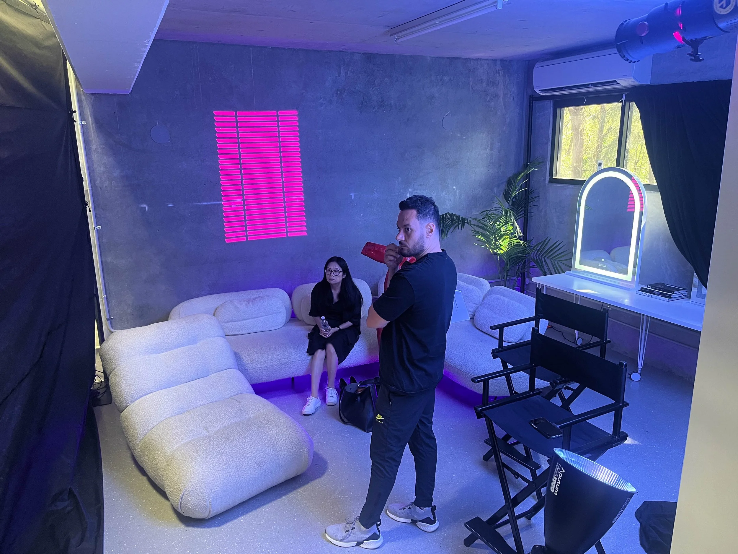 People in a dimly lit room with a neon pink light display on a gray wall, some sitting on beige couches, and a man in black standing while sipping from a red cup. There is a woman with glasses sitting on the couch and a large mirror with LED lights on a table near a window with a view of green trees outside.