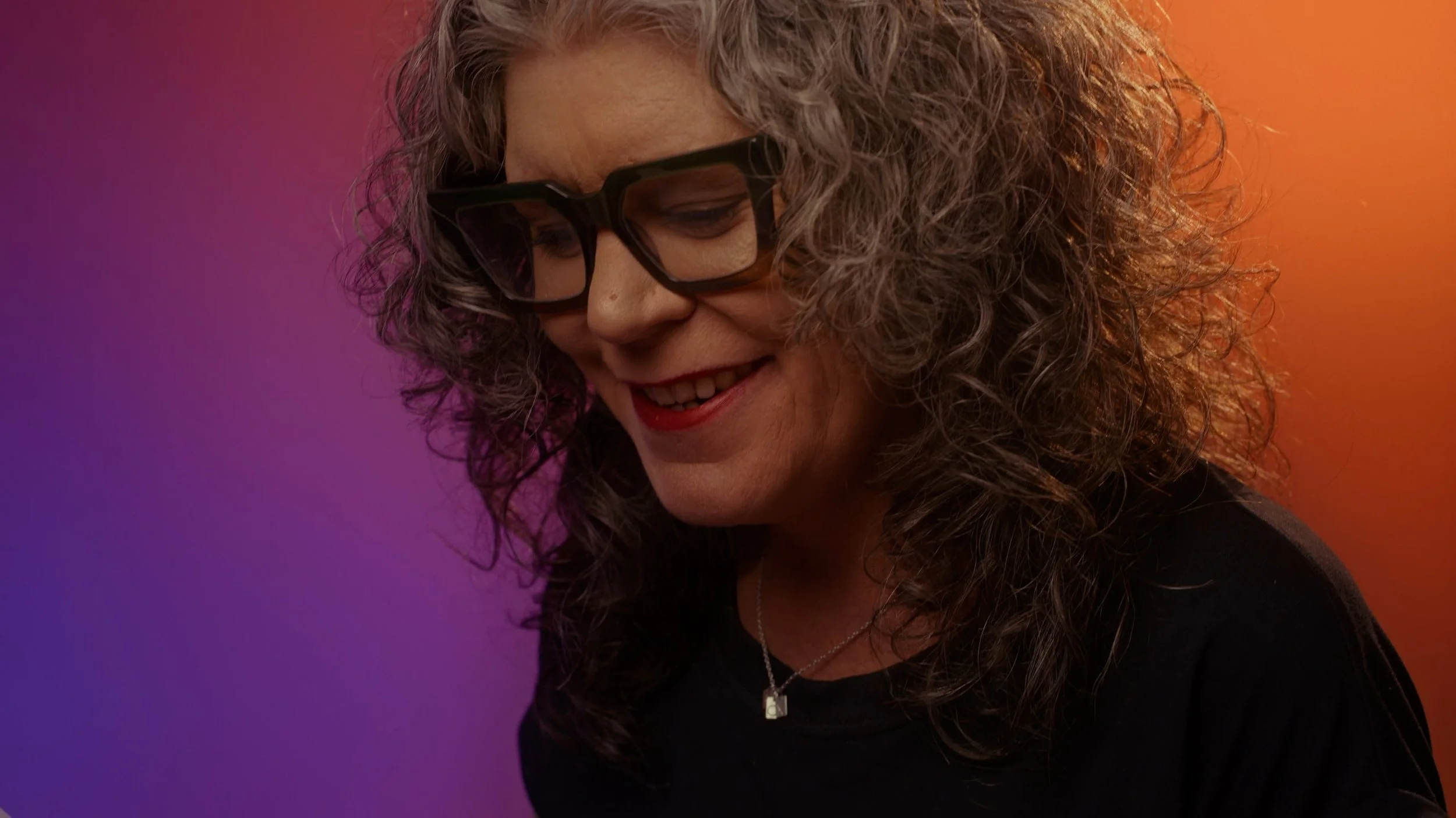A smiling woman with curly gray hair wearing black glasses and a black shirt, against a colorful gradient background.