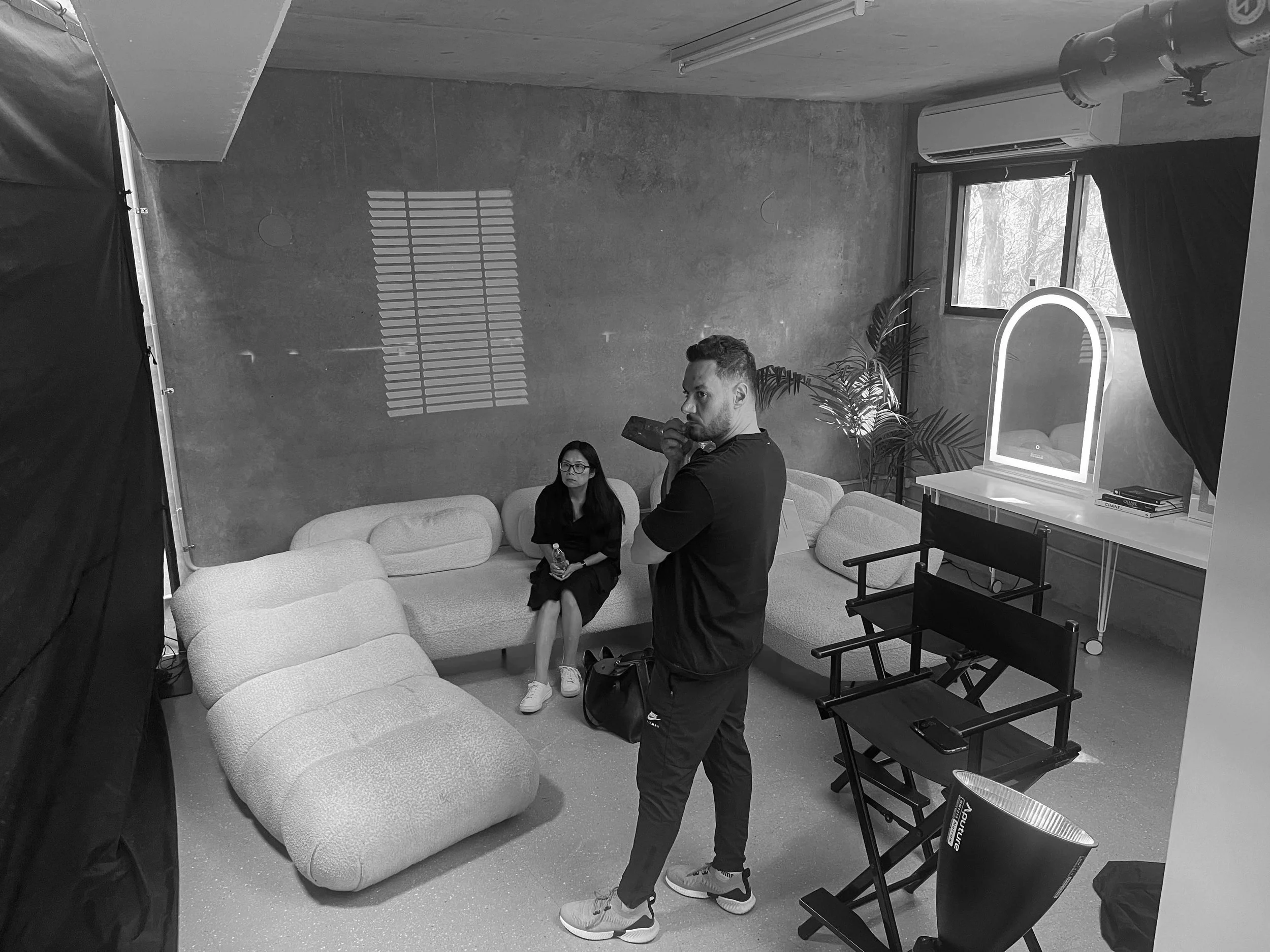 A black and white photo of a woman sitting on a curved sofa holding a bottle, a man standing nearby drinking from a bottle, in a room with minimal decor and a large window with sunlight casting shadows on the wall.