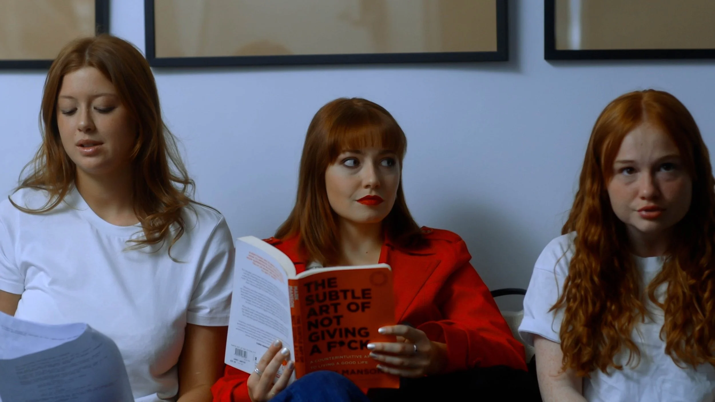Three women sitting on a bench against a white wall with framed pictures. The woman on the left has long reddish hair, is wearing a white shirt, and is looking down. The woman in the middle has shoulder-length red hair, is wearing a red jacket, and is holding a book titled "The Subtle Art of Not Giving a F*ck." The woman on the right has long curly red hair and is wearing a white shirt, looking slightly to the side.
