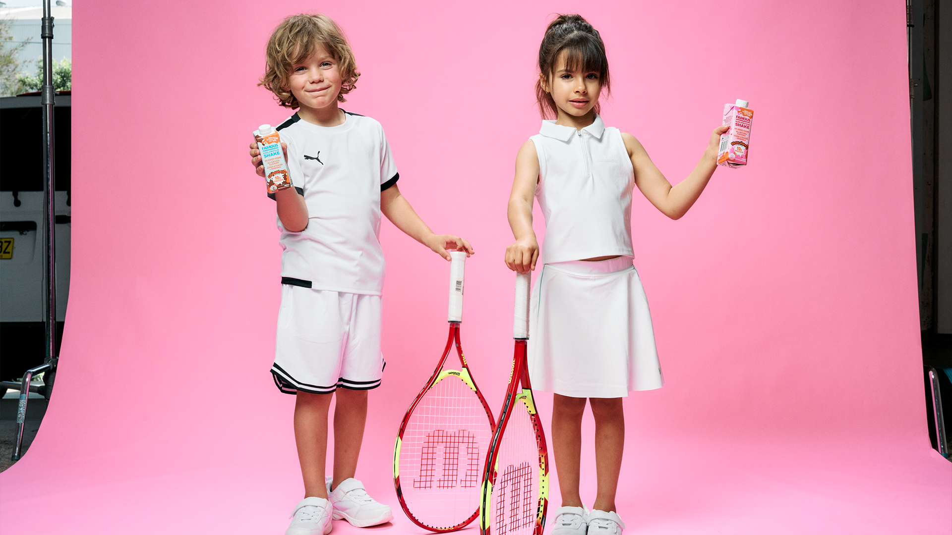 Two children, a boy and a girl, dressed in tennis outfits, standing in front of a pink backdrop, holding tennis rackets and cartons of chocolate milk.