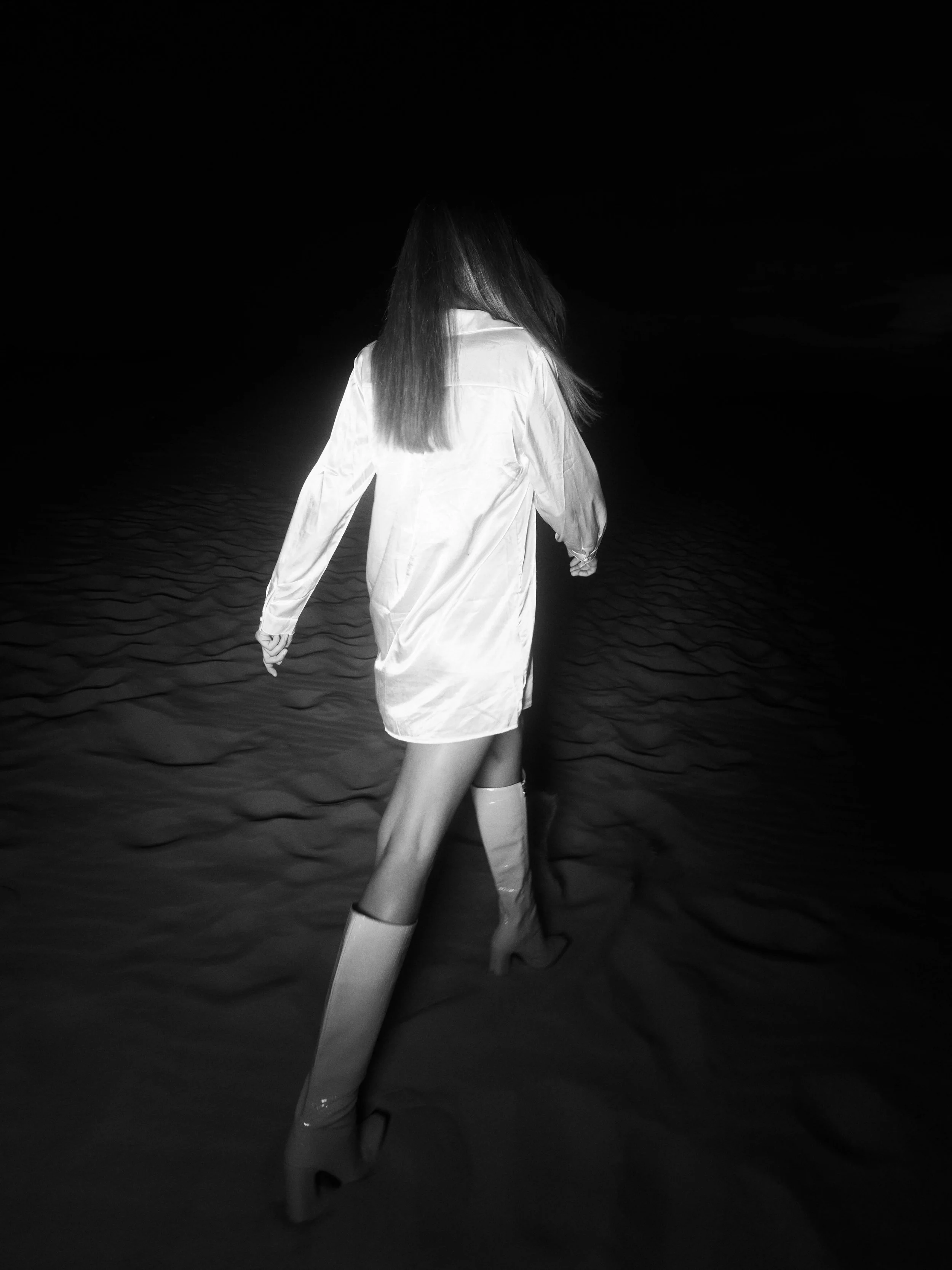 A woman in a white coat and high boots walking on sand at night.