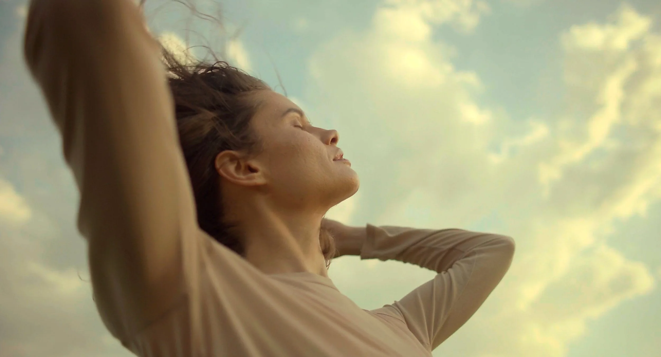 A woman with closed eyes and arms behind her head outdoors during the daytime with a cloudy sky in the background.
