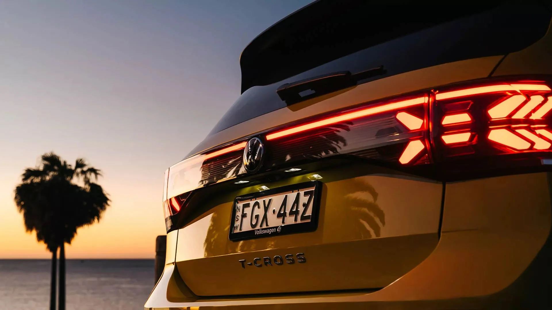 Close-up of the rear of a yellow Volkswagen T-Cross with tail lights illuminated during sunset, with a silhouetted palm tree and ocean in the background