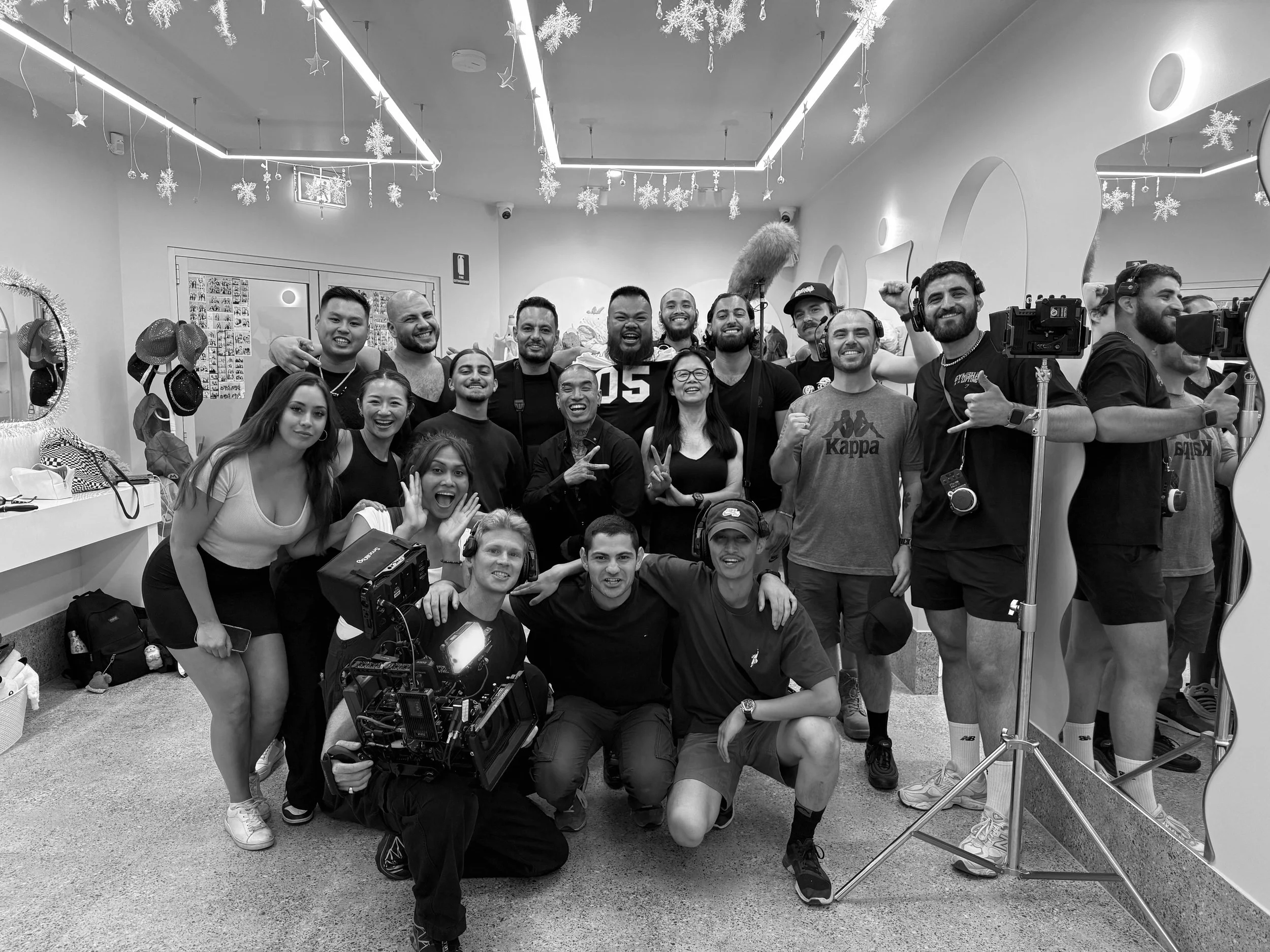 A group of people on a film set smiling and posing for a photo, with some wearing headsets and holding cameras, in a decorated indoor setting with hanging snowflakes and festive decor.