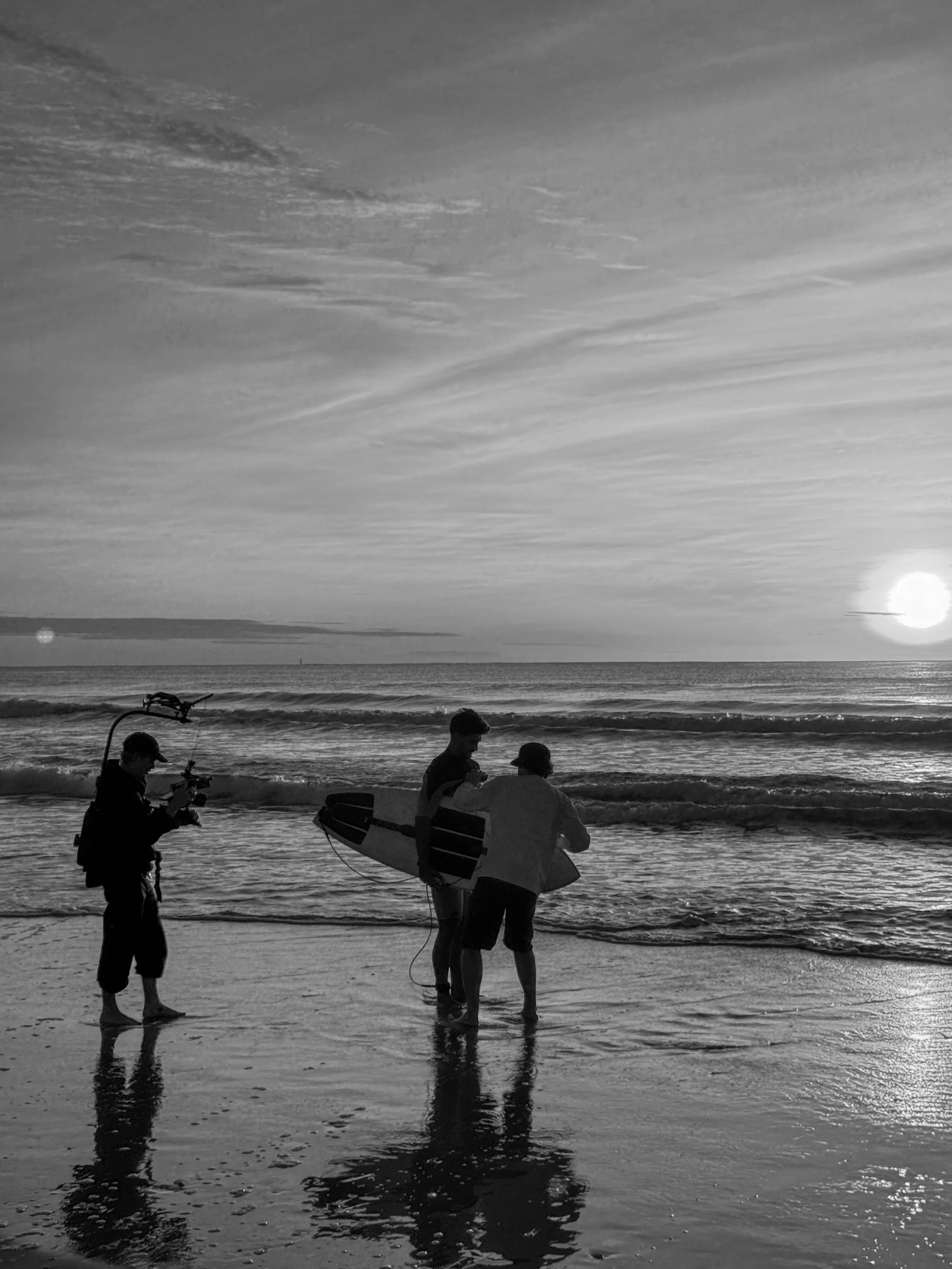People at the beach during sunset, some holding a surfboard and others standing in the water, with the sun near the horizon and sky with wispy clouds.
