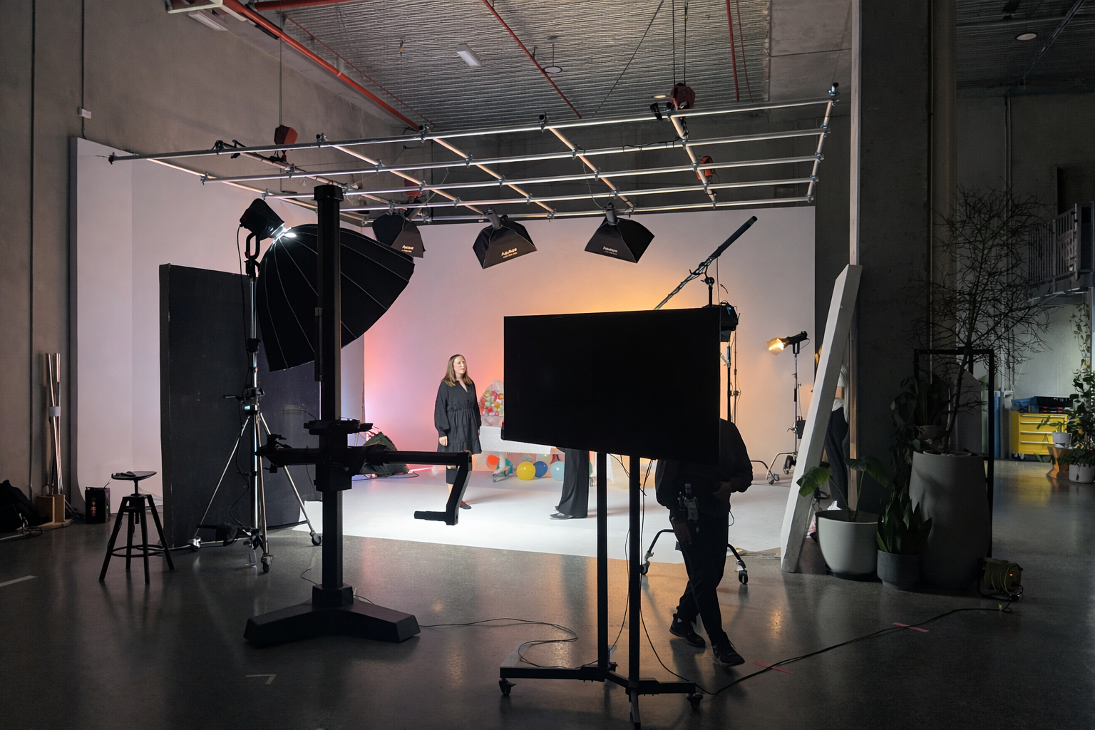Photo of a professional photo studio setup with lighting, a backdrop, and a woman in a black dress standing on a white floor in front of colorful balloons and background decorations.