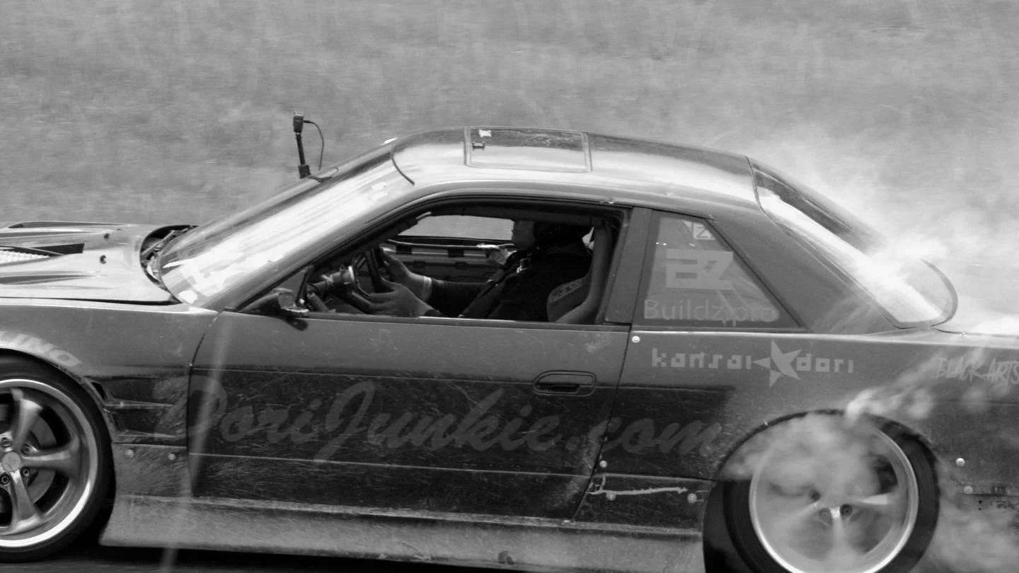 A black and white photo of a race car in motion during a drift, with smoke coming from the rear tires, and a driver inside wearing a helmet.
