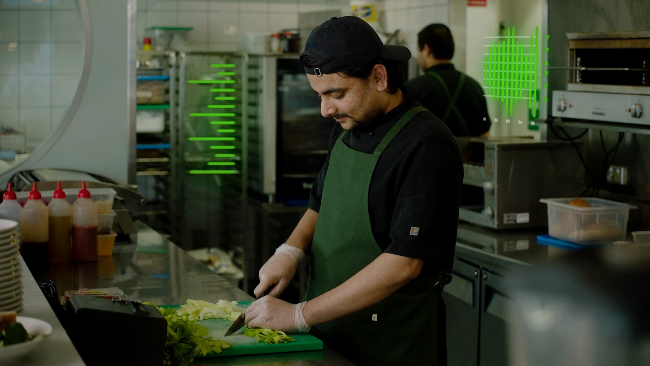 Male chef wearing a black cap, black shirt, and green apron chopping lettuce in a restaurant kitchen with cooking equipment and a worker in the background.