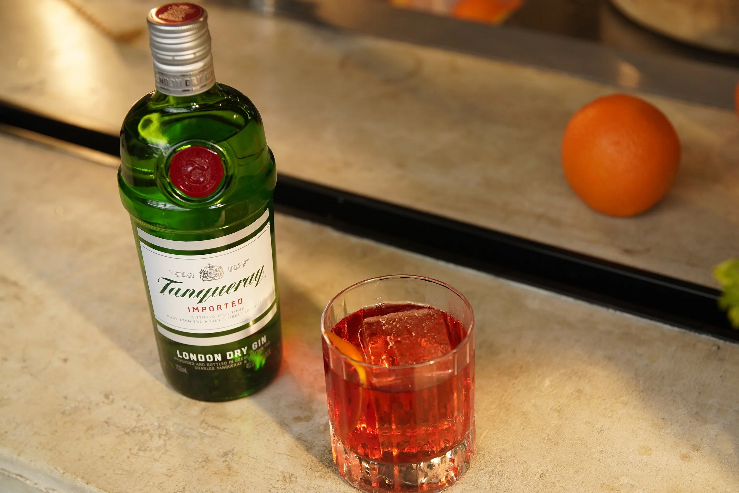 A bottle of Tanqueray London Dry Gin and a glass filled with red liquid and ice cubes on a kitchen counter, with a blurred orange fruit in the background.