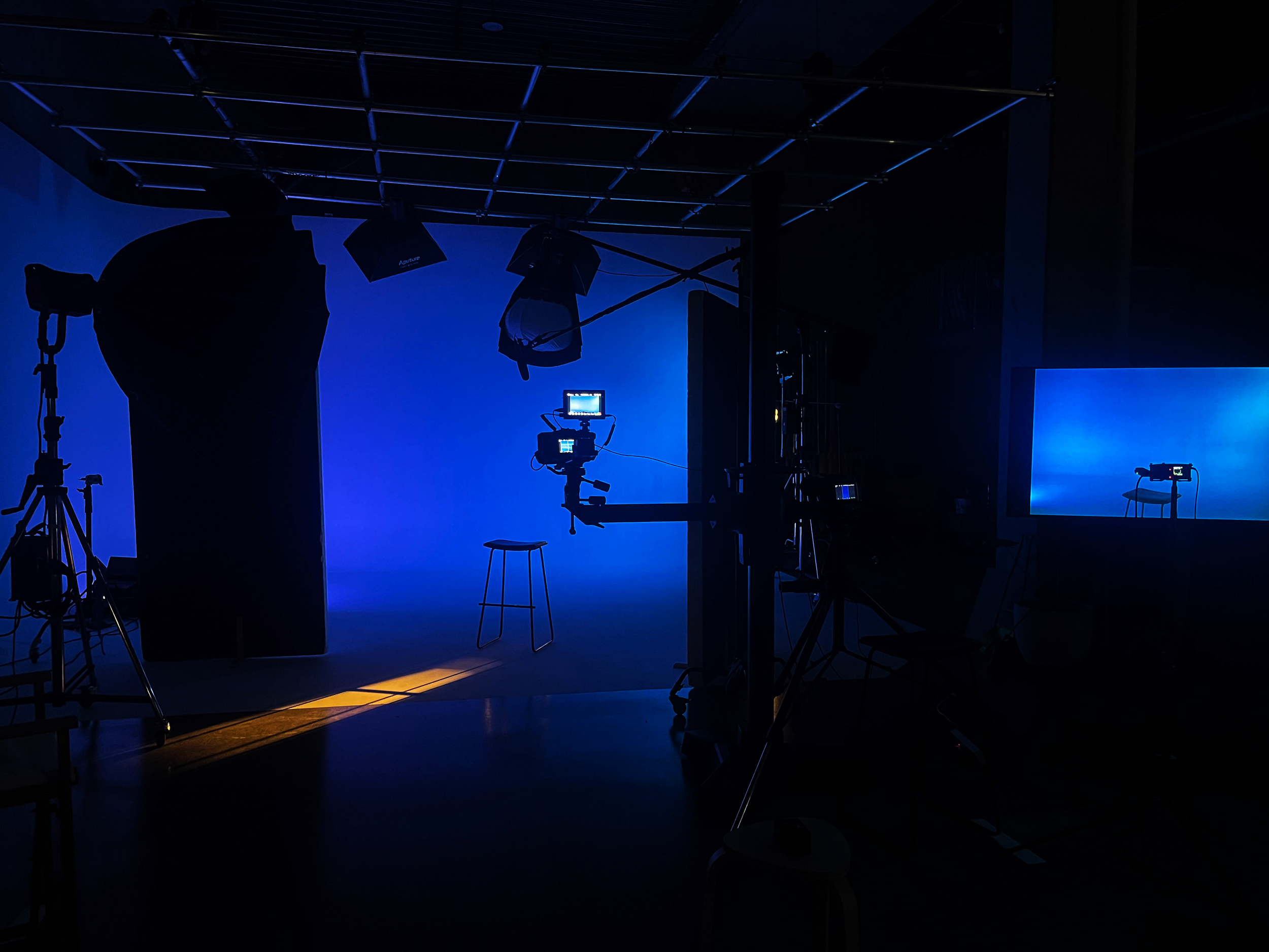 A dimly lit photography studio with blue lighting, various studio equipment, lights, camera, and a stool in front of a blue backdrop.