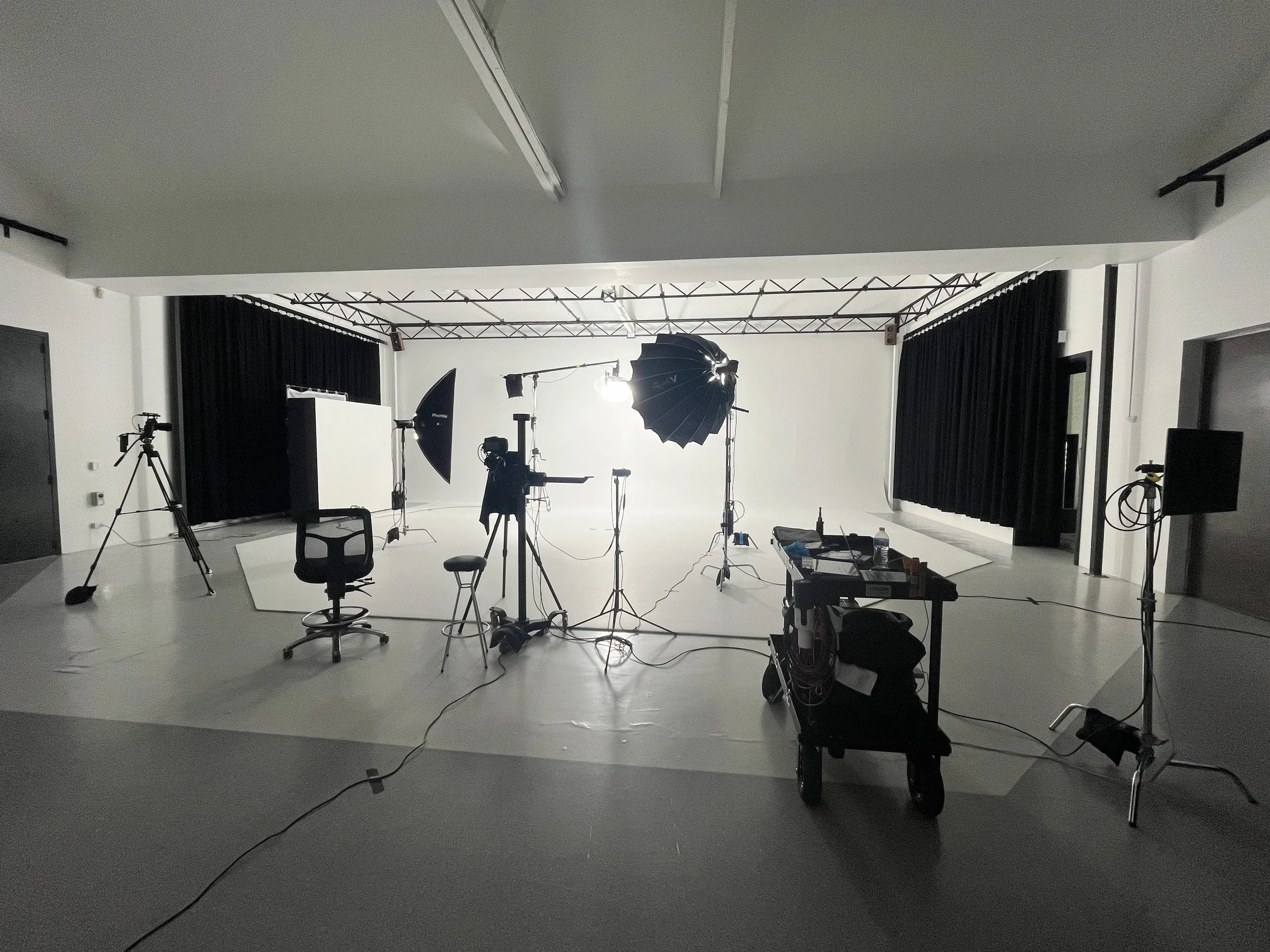 A photography studio with lighting equipment, cameras, and tripods set up, white backdrop, and black curtains on sides.