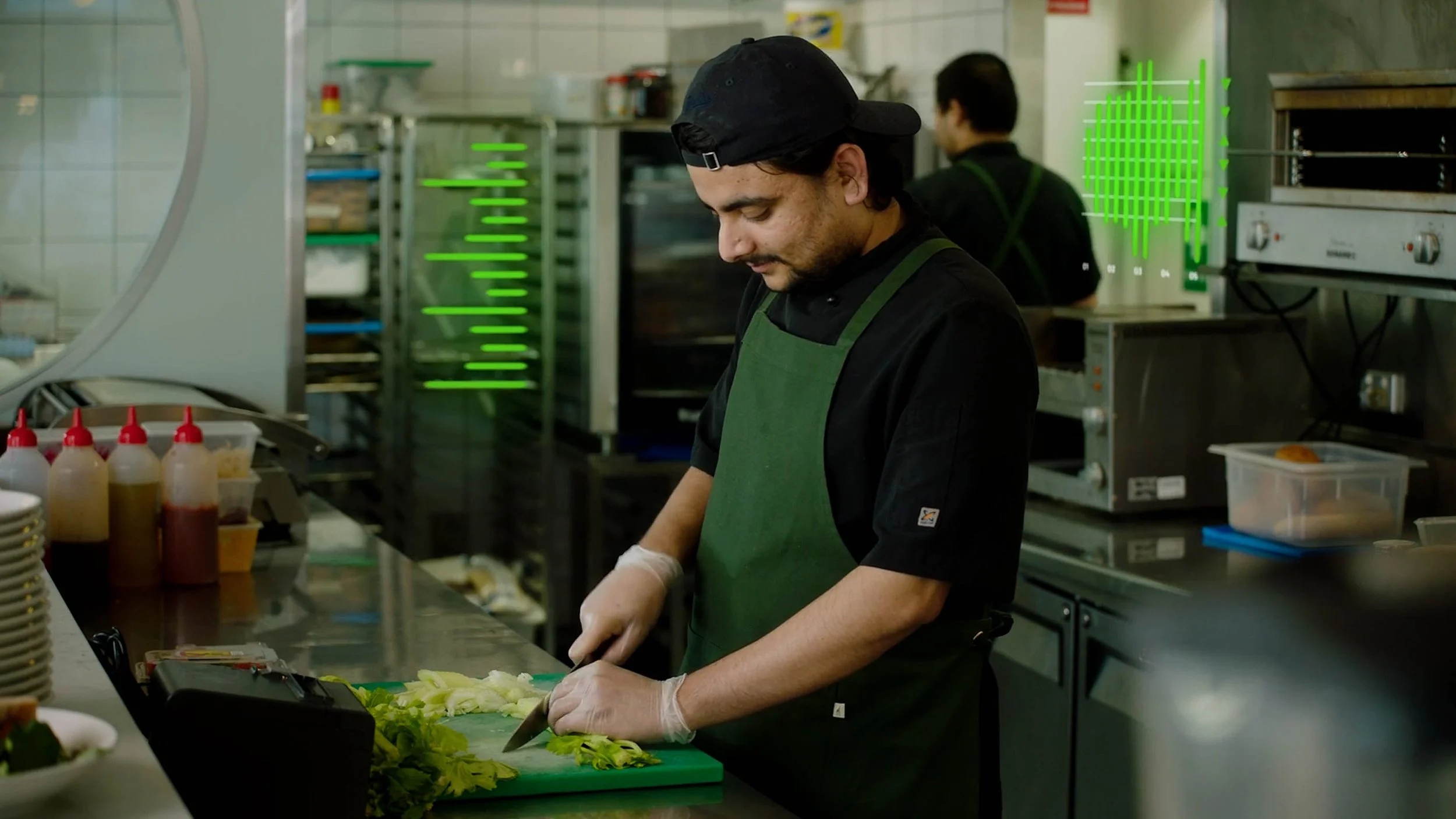 A man wearing a black cap, black shirt, and green apron chopping vegetables in a commercial kitchen.