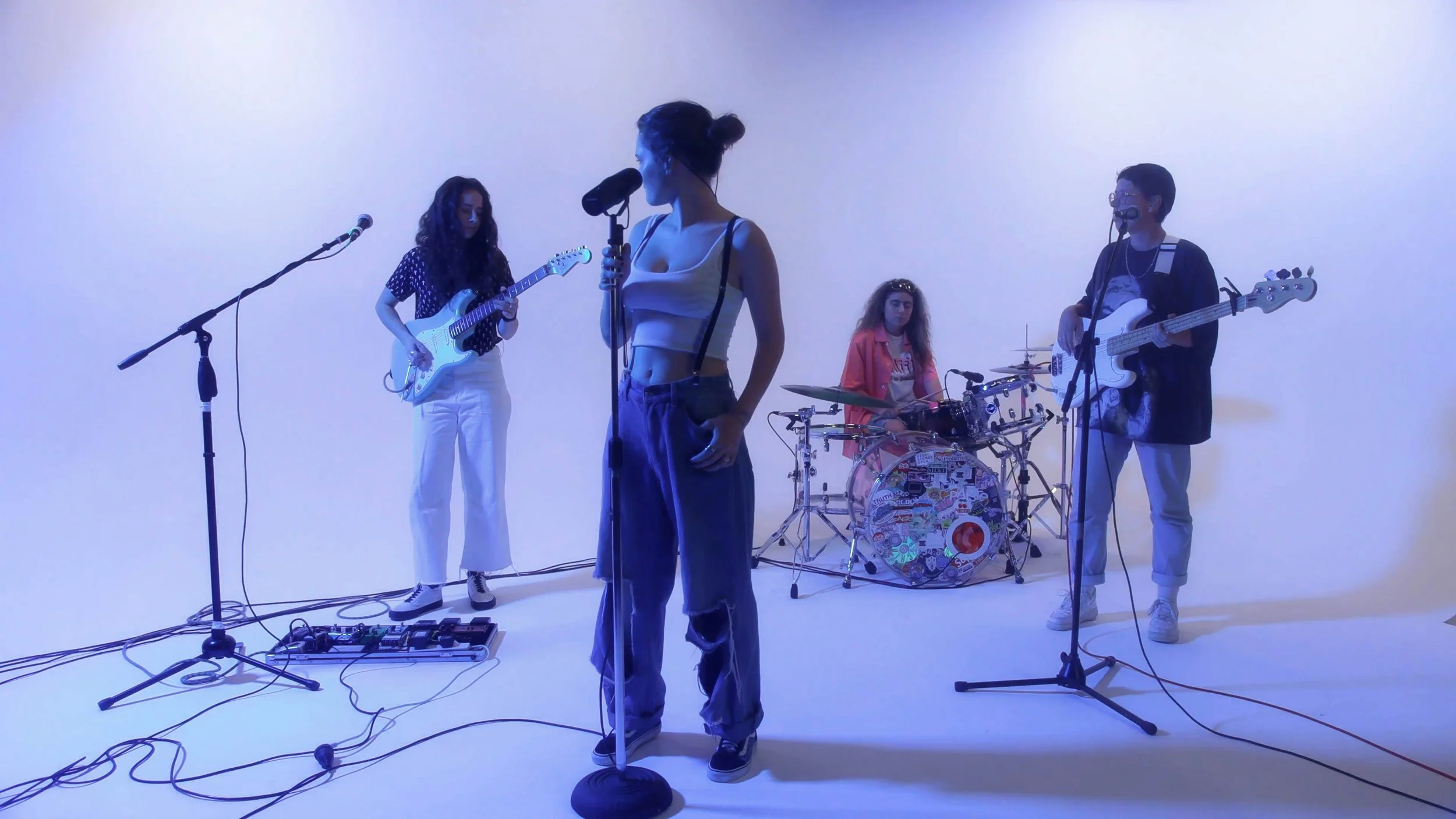 A band of five women performing with musical instruments on a white studio background, illuminated with blue lighting.