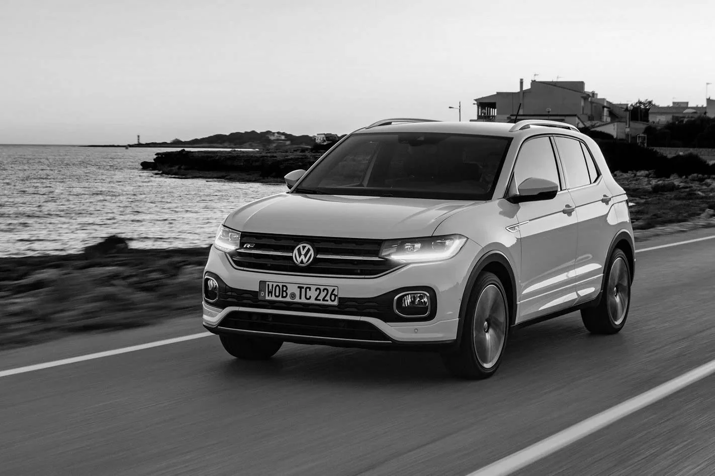 A Volkswagen SUV driving along a coastal road with houses and a lighthouse in the background