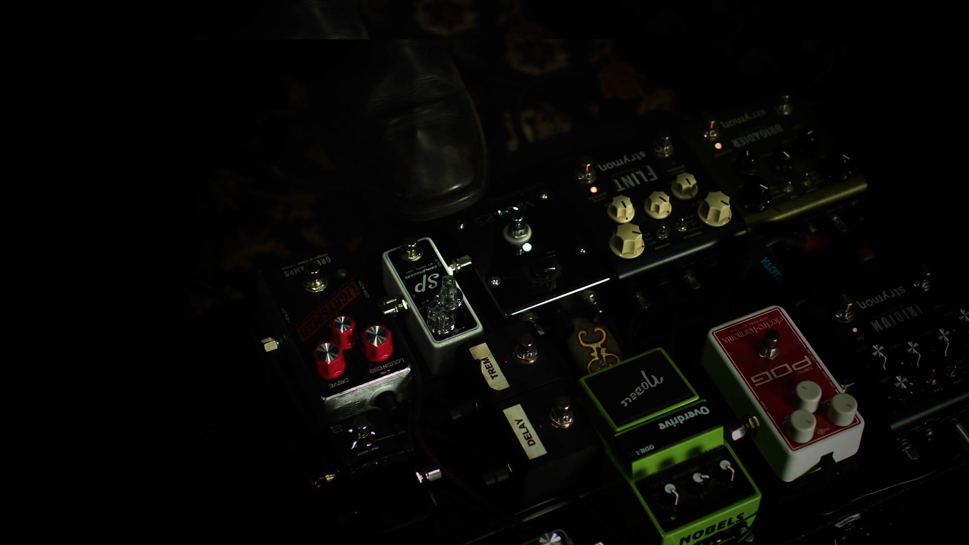Close-up of a guitar effects pedalboard with various pedals, including distortion, delay, and modulation effects, under dim lighting.