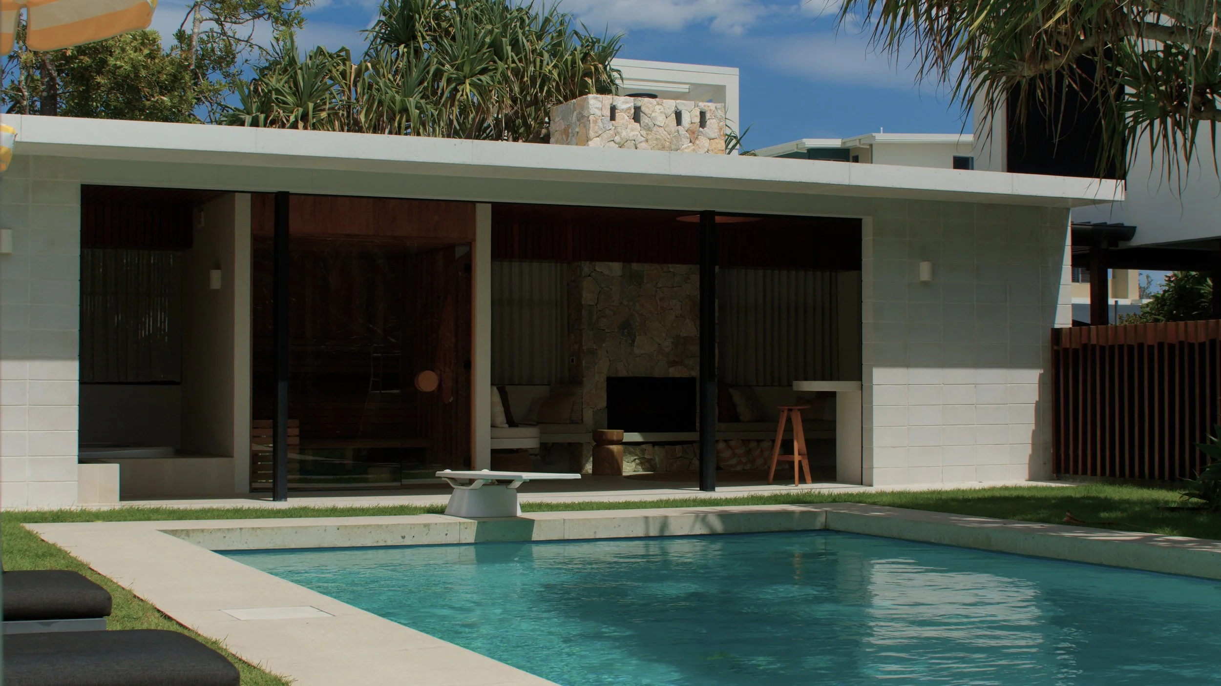 Modern house with a swimming pool in the backyard, glass sliding doors, and outdoor furniture under a flat roof, with a blue sky and trees in the background.