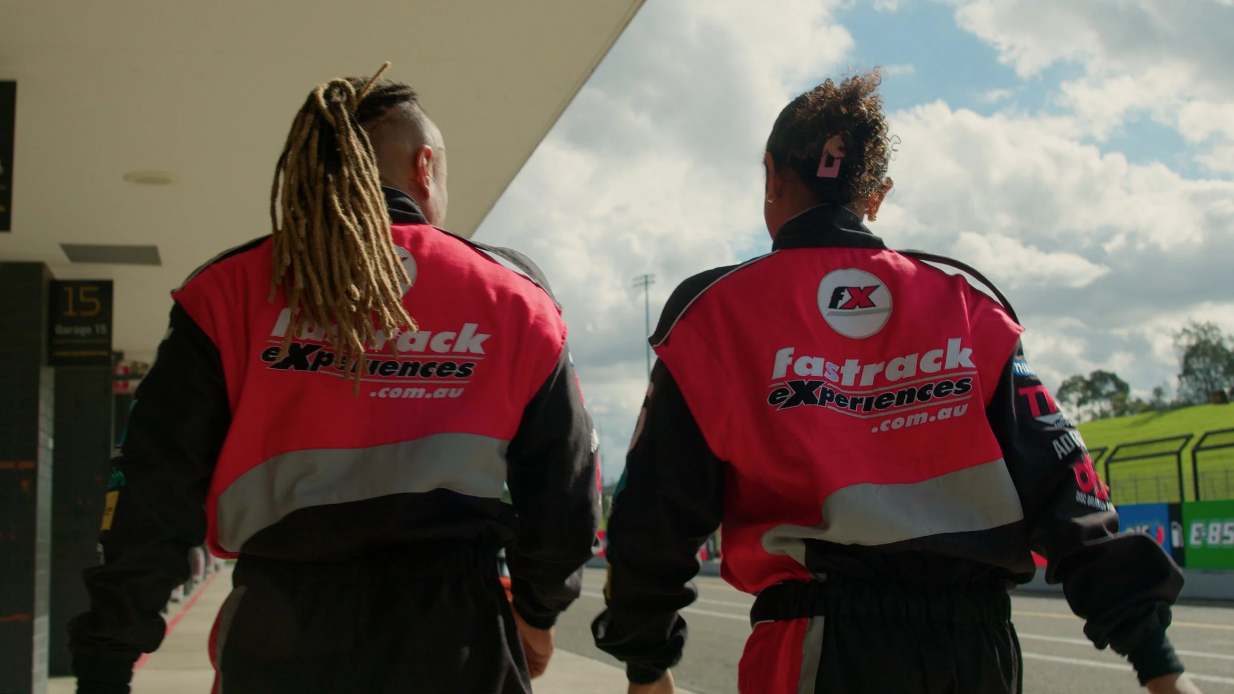 Two race car drivers wearing red and black FastTrack eXperiences jackets walking away from the camera at a race track.