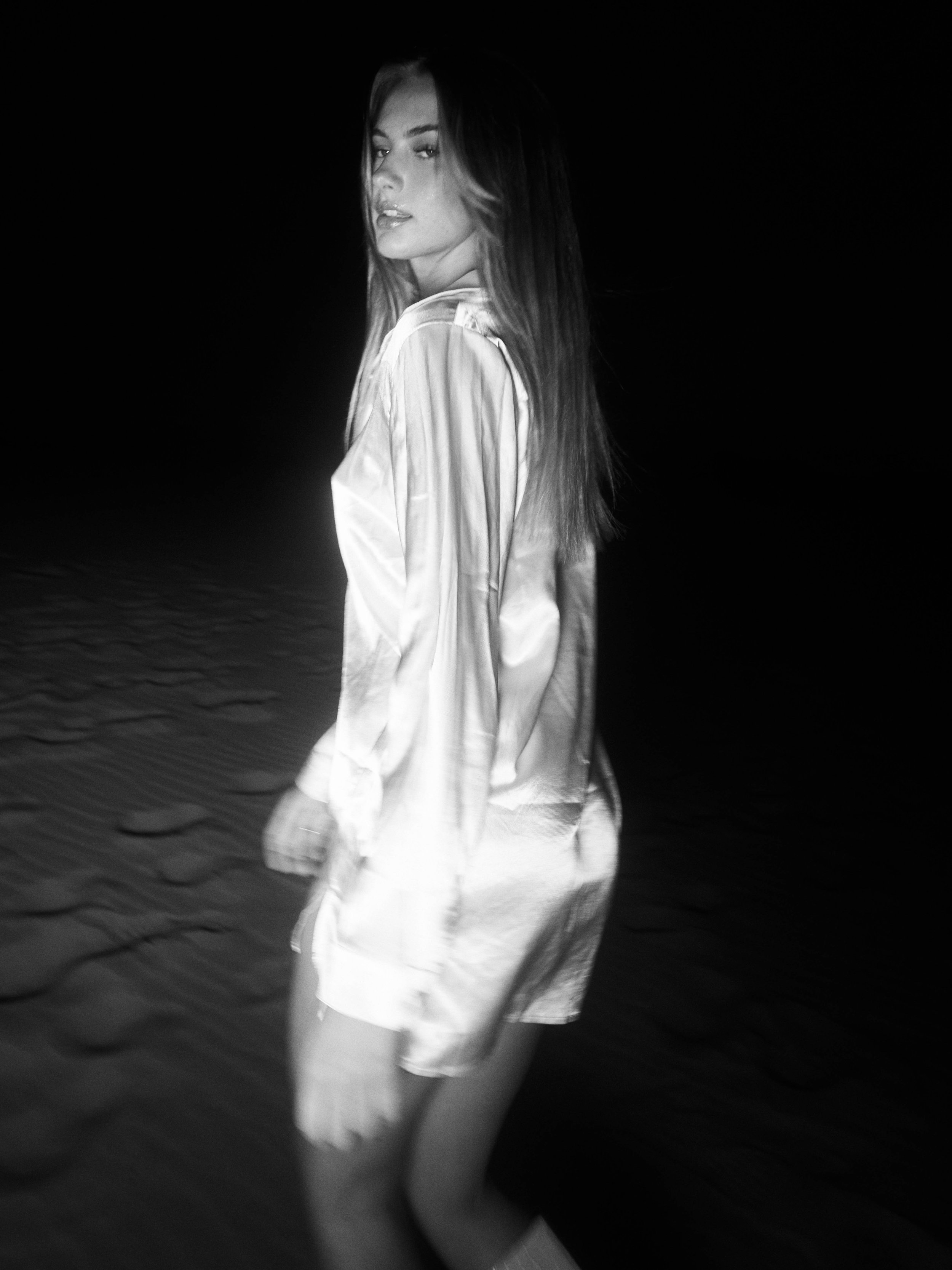 A woman in a silk robe stands on the beach at night, illuminated by a bright light against a dark background.