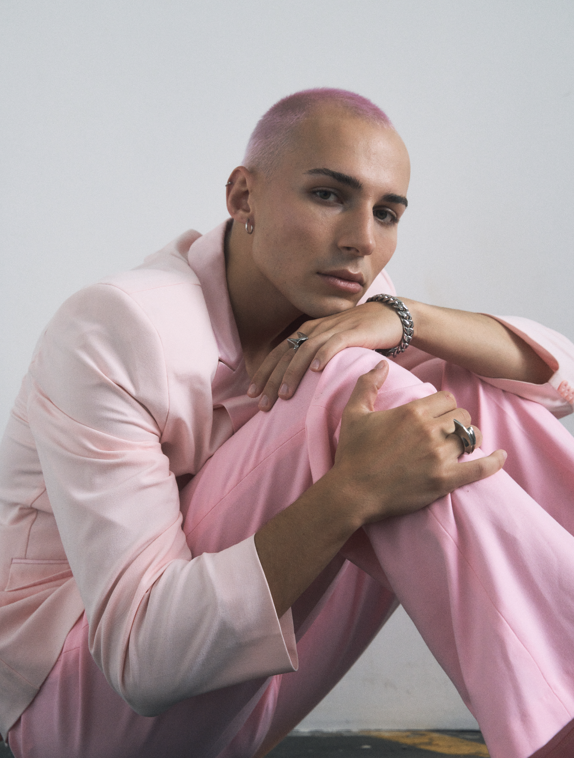 A person with pink hair and a pink suit leaning forward with arms crossed over their knees, posing against a plain background.