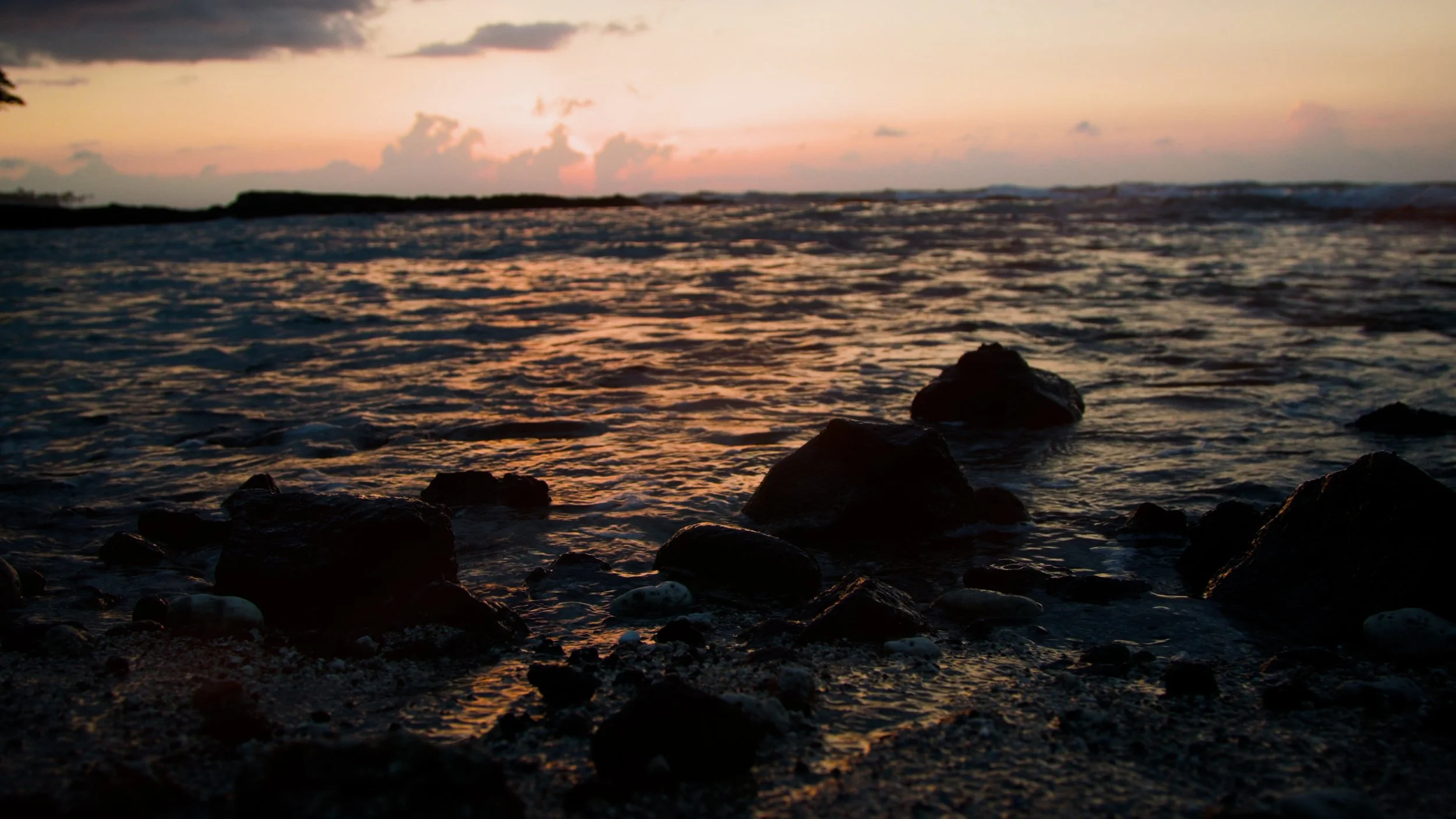 A view of the ocean at sunset with waves and rocks in the foreground and cloudy sky in the background.