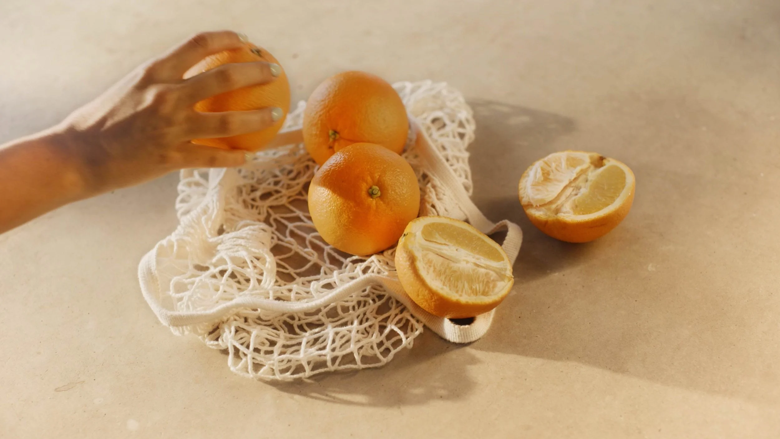 A hand reaching to pick up an orange among three oranges on a white mesh bag and two halved oranges on a beige surface.