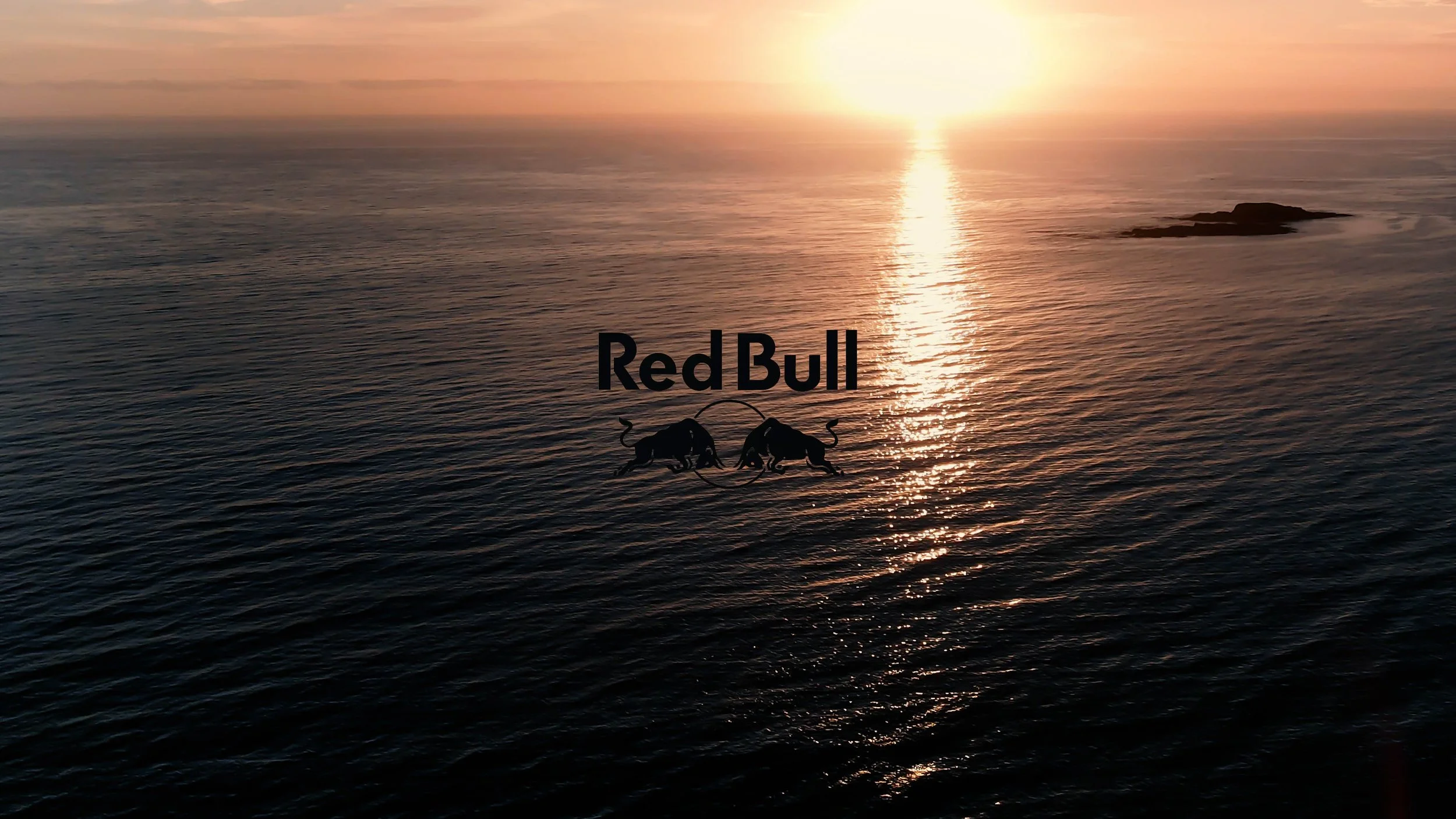 Sunset over the ocean with gentle waves and rocks, featuring a Red Bull logo with two bulls and directional text.