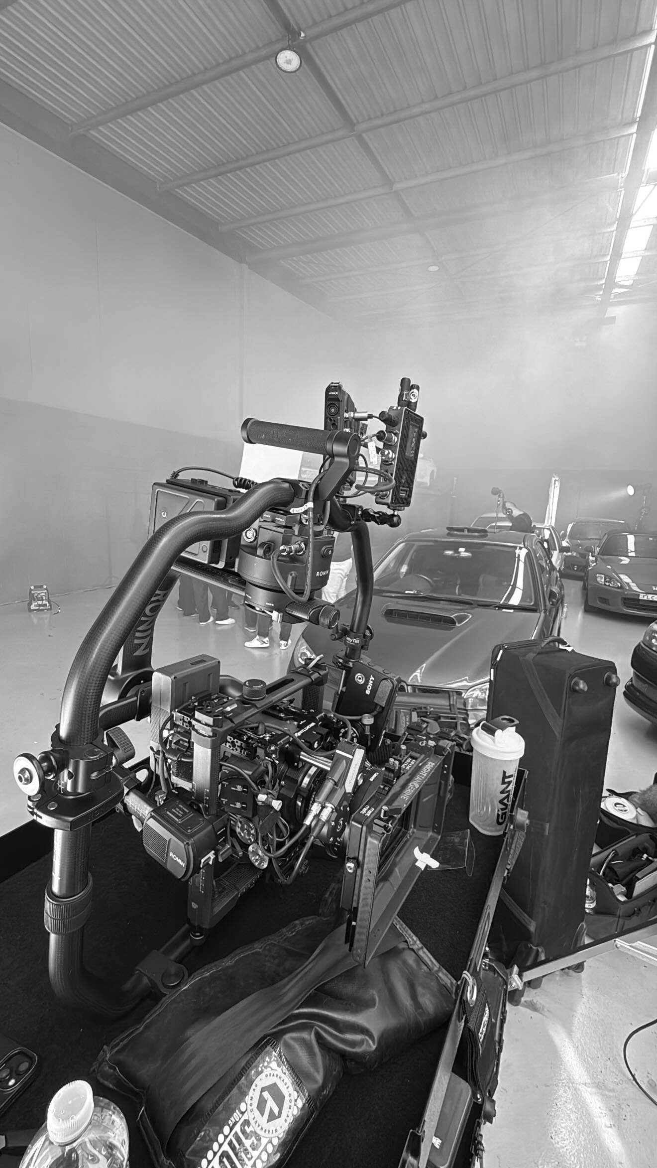 Behind the scenes of a film or TV production set with a camera rig in the foreground, multiple parked cars in the background, and crew members partially visible, all in a large indoor warehouse or studio space.