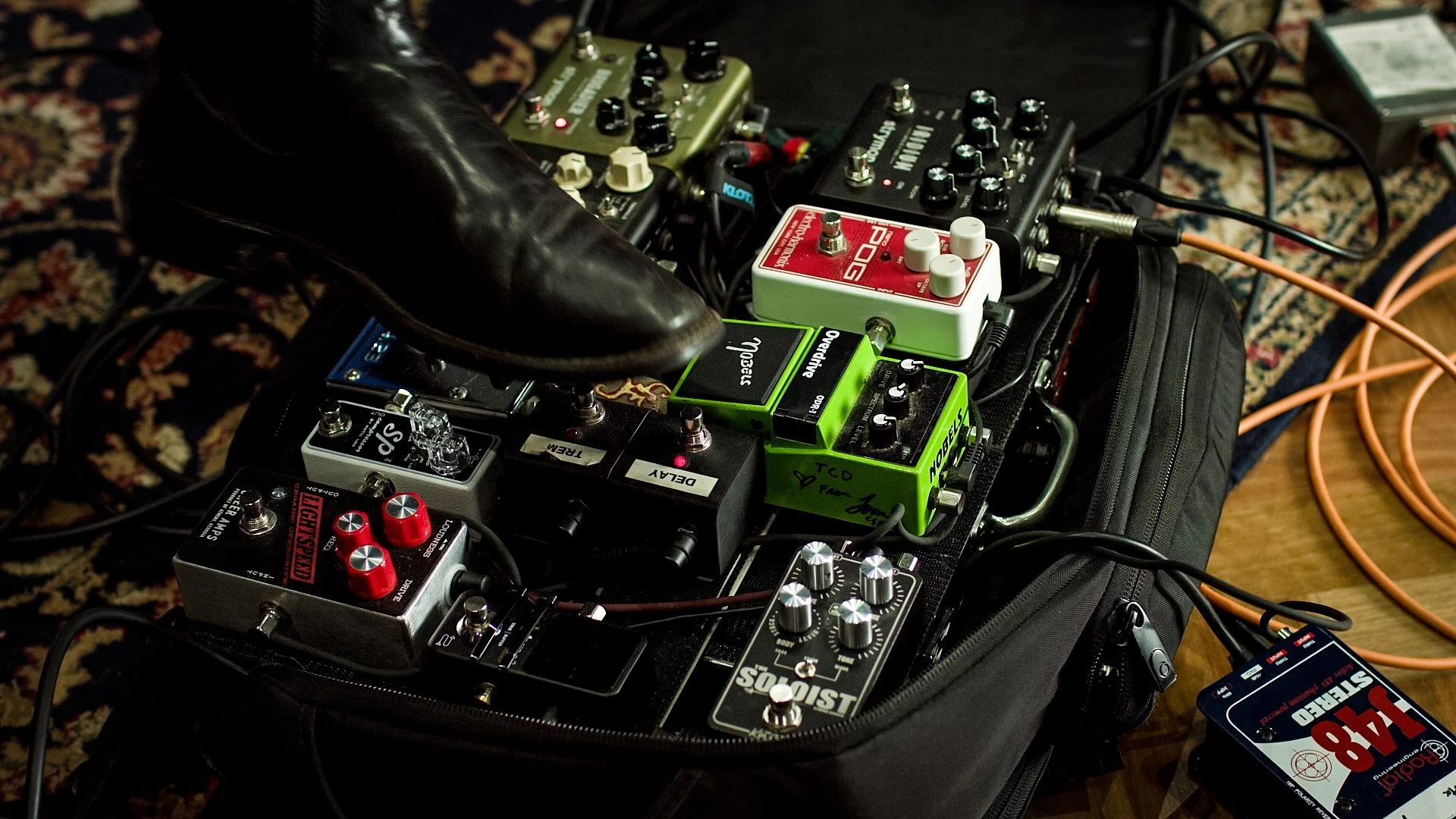 Multiple guitar effects pedals connected with cables on a pedalboard, with a black carrying case underneath, on a patterned carpet.