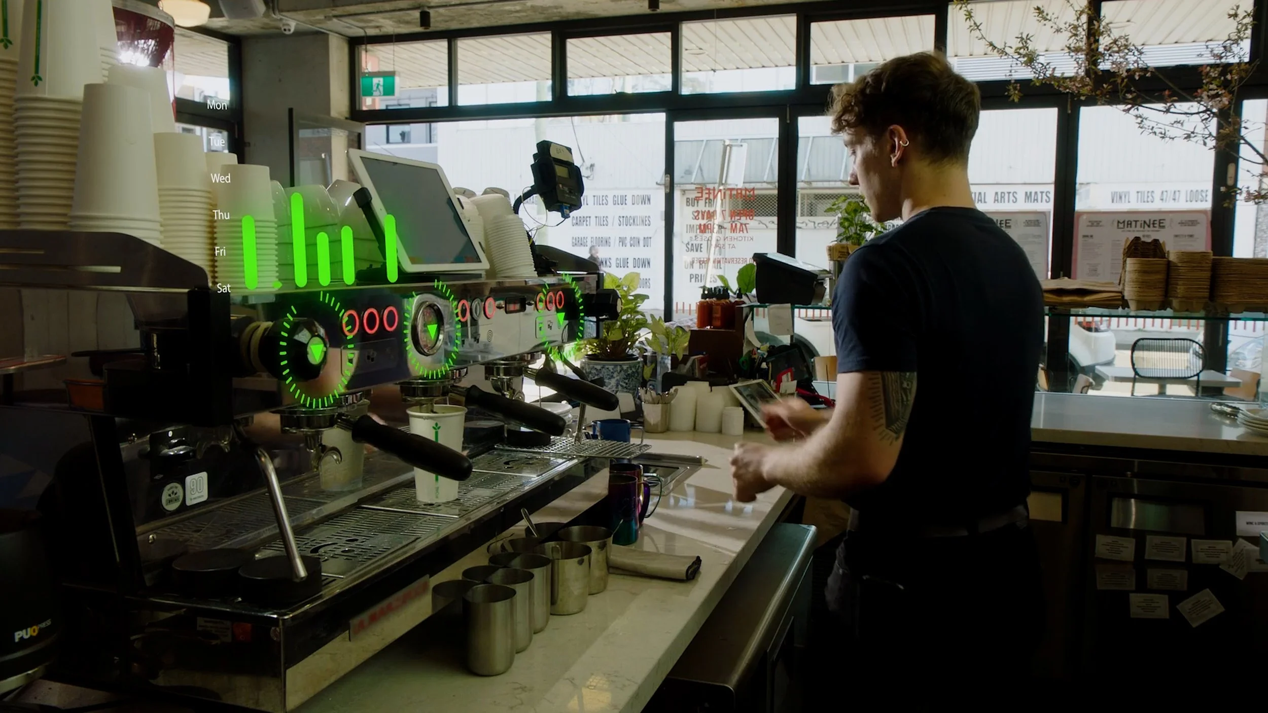 Barista in a coffee shop preparing drinks behind the counter, with a coffee espresso machine, cups, and a digital display overlay showing a weekly schedule.