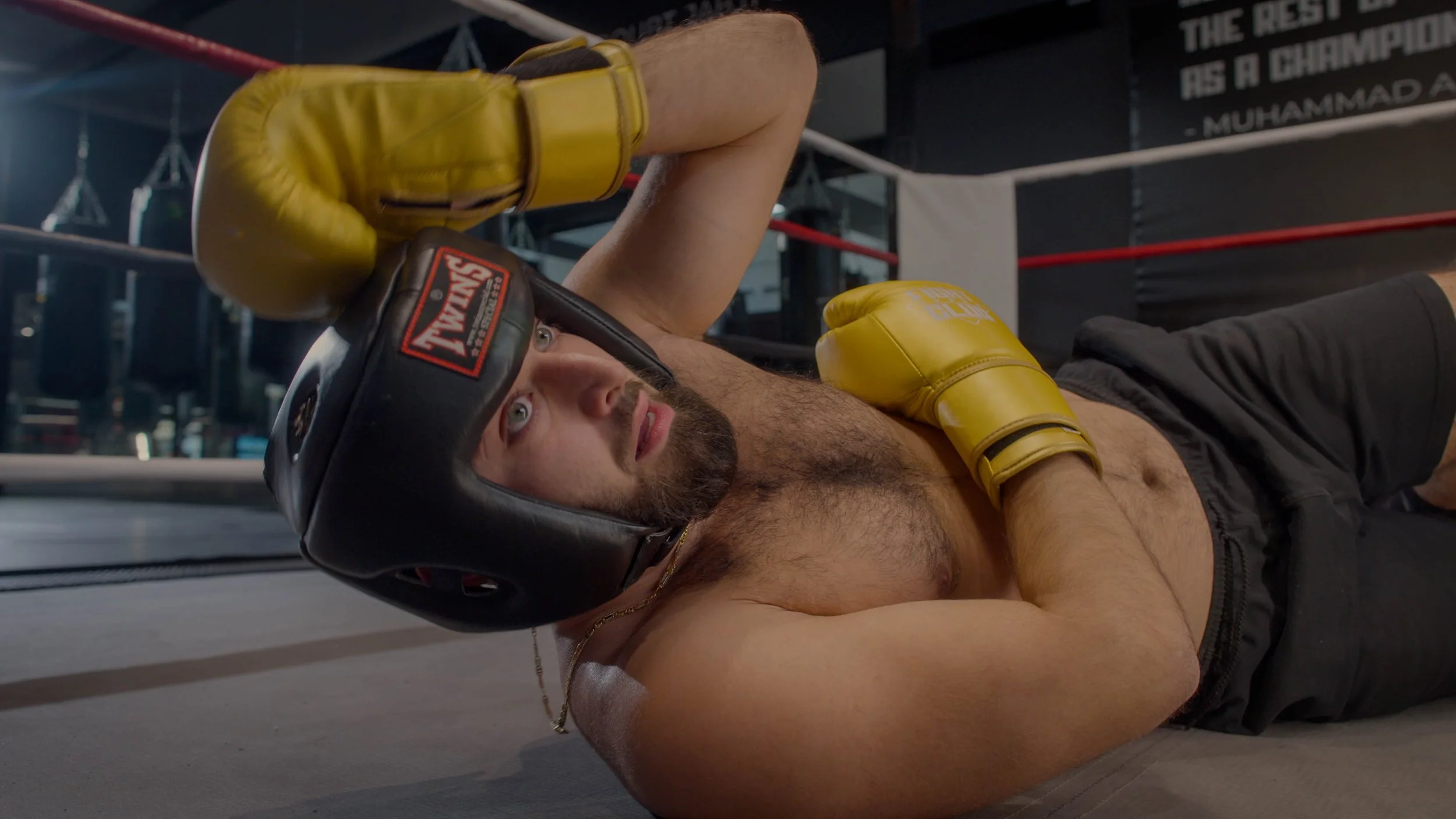 A male boxer lying on the floor inside a boxing ring, wearing a black headgear and yellow boxing gloves, looking up toward the ceiling.