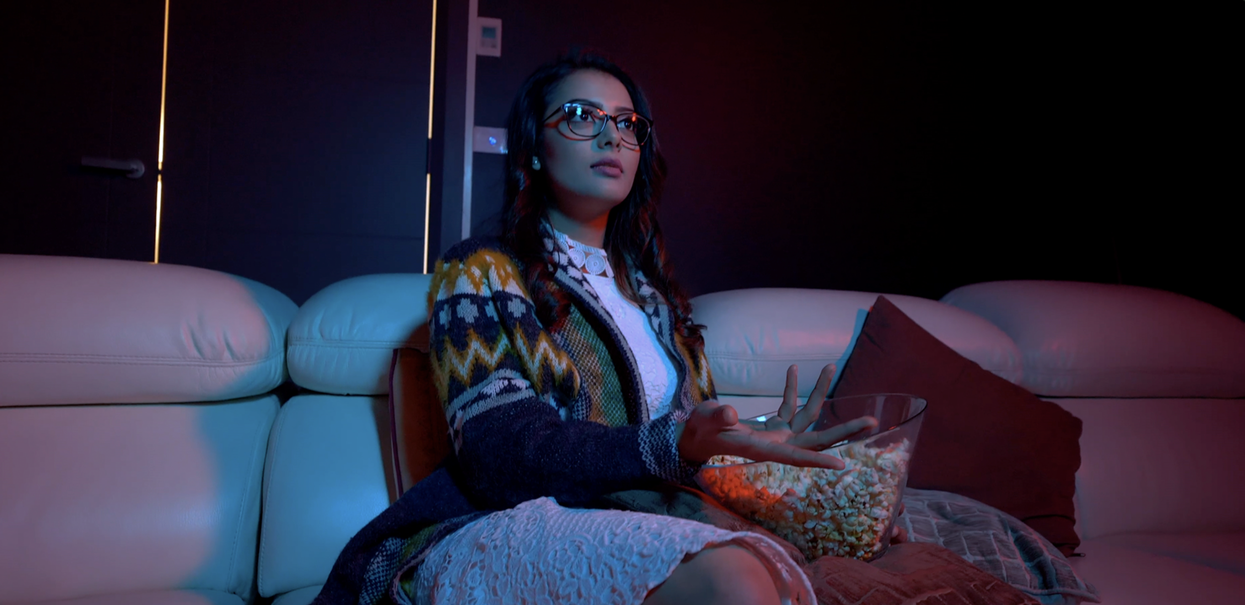 A woman with dark hair, glasses, and a colorful sweater sitting on a cream-colored sofa, illuminated by blue and pink lighting in a modern room, holding a bowl of popcorn.