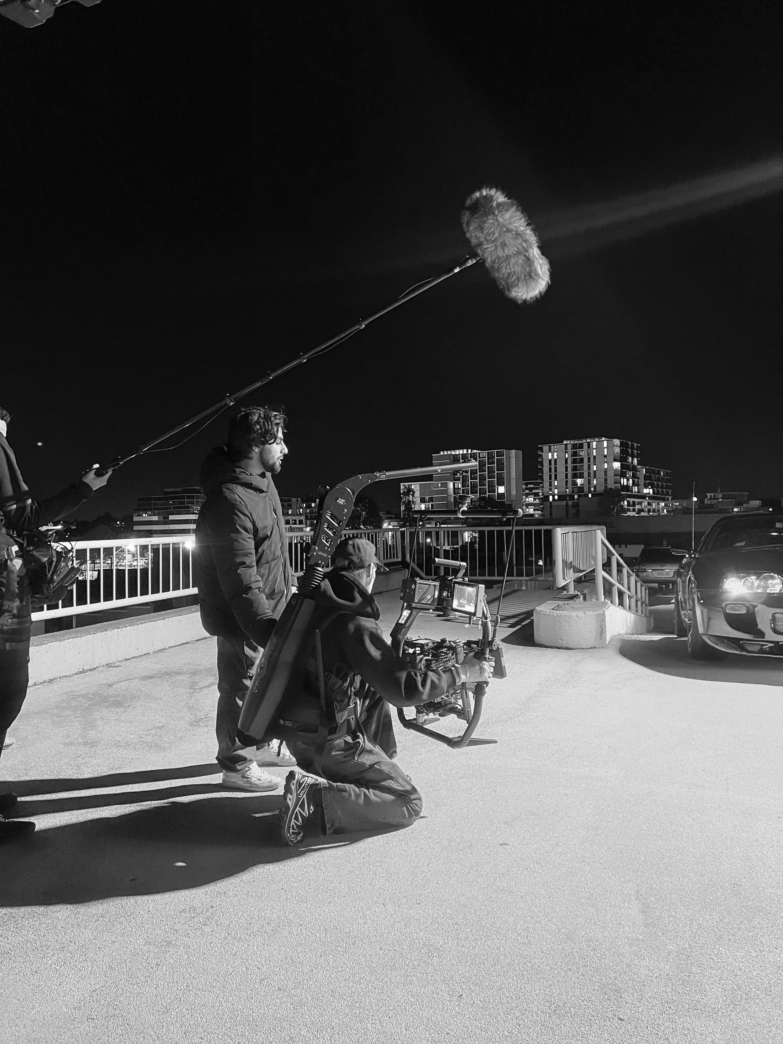 Black and white photo of a film crew filming at night on a parking lot rooftop. One person kneels with a camera rig, others stand nearby. A boom microphone is held above them.