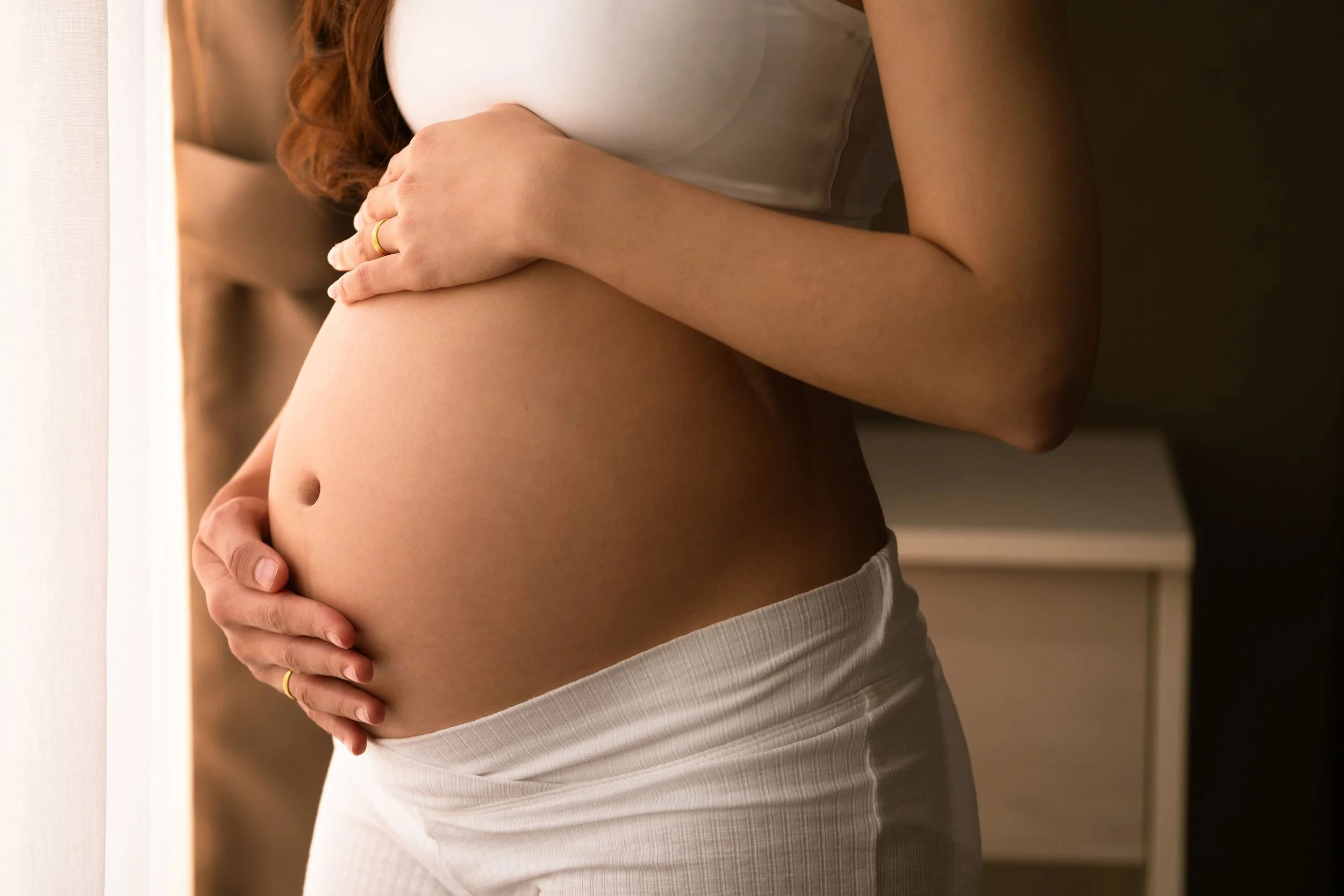 A pregnant woman gently cradles her belly with both hands, standing near a window with natural light illuminating her abdomen.
