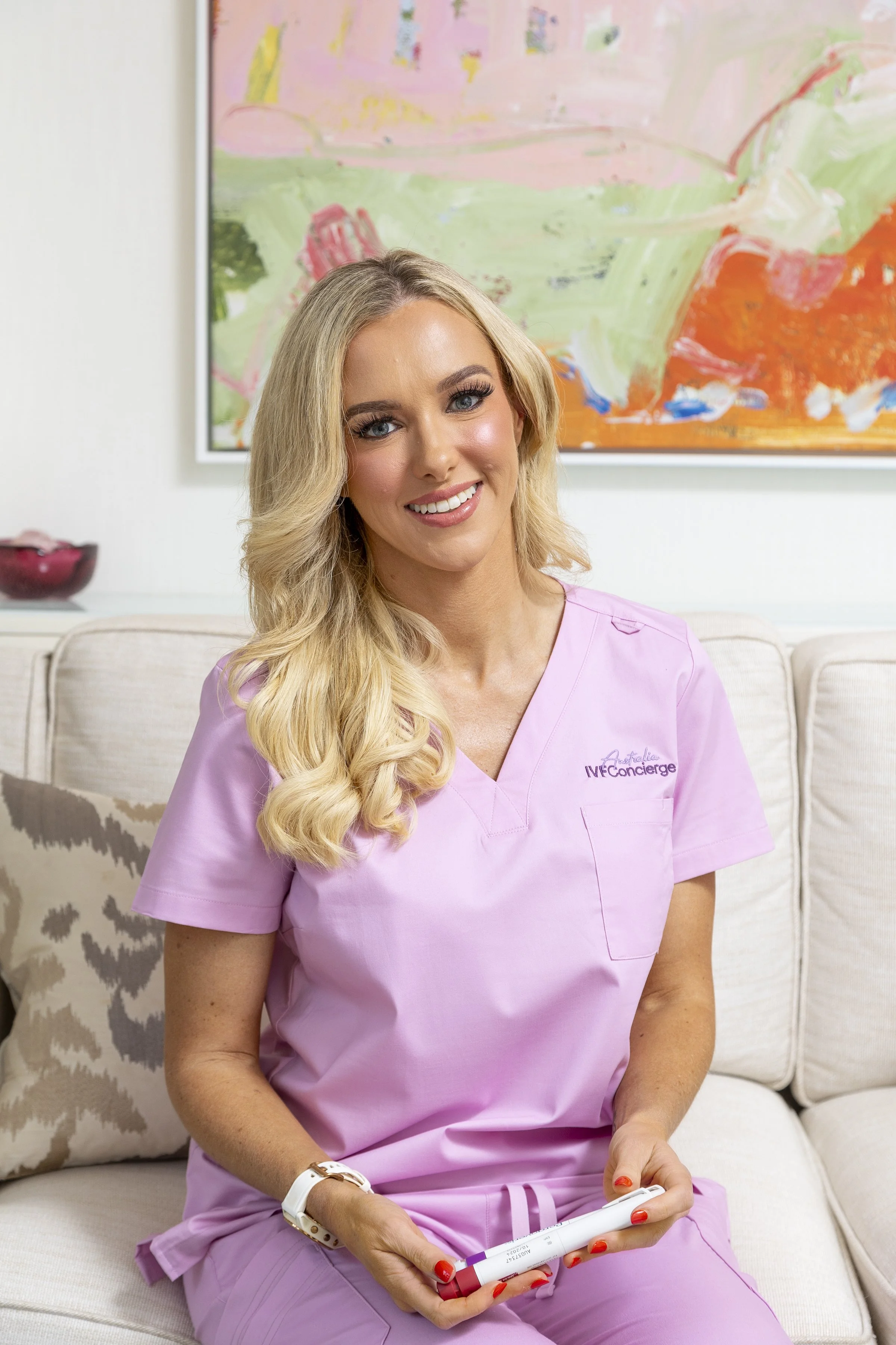 A smiling woman in pink scrubs sitting on a sofa, holding a pregnancy test. Behind her is abstract artwork and a patterned pillow.