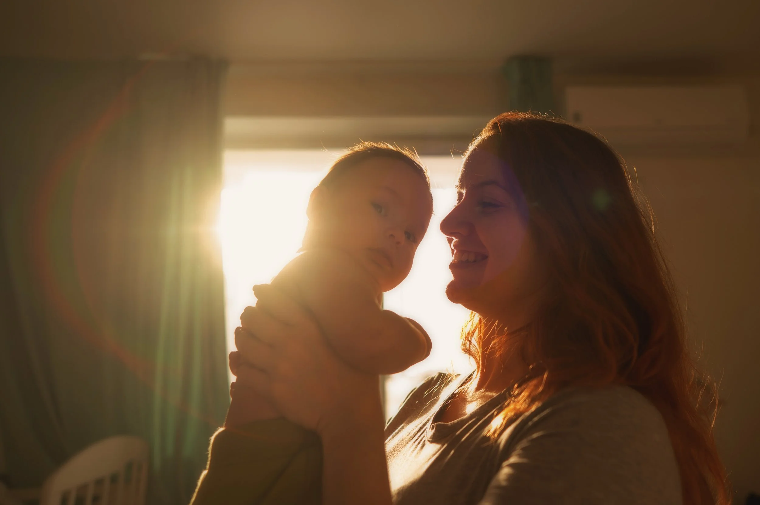 A woman holding a baby near a bright window during sunset.