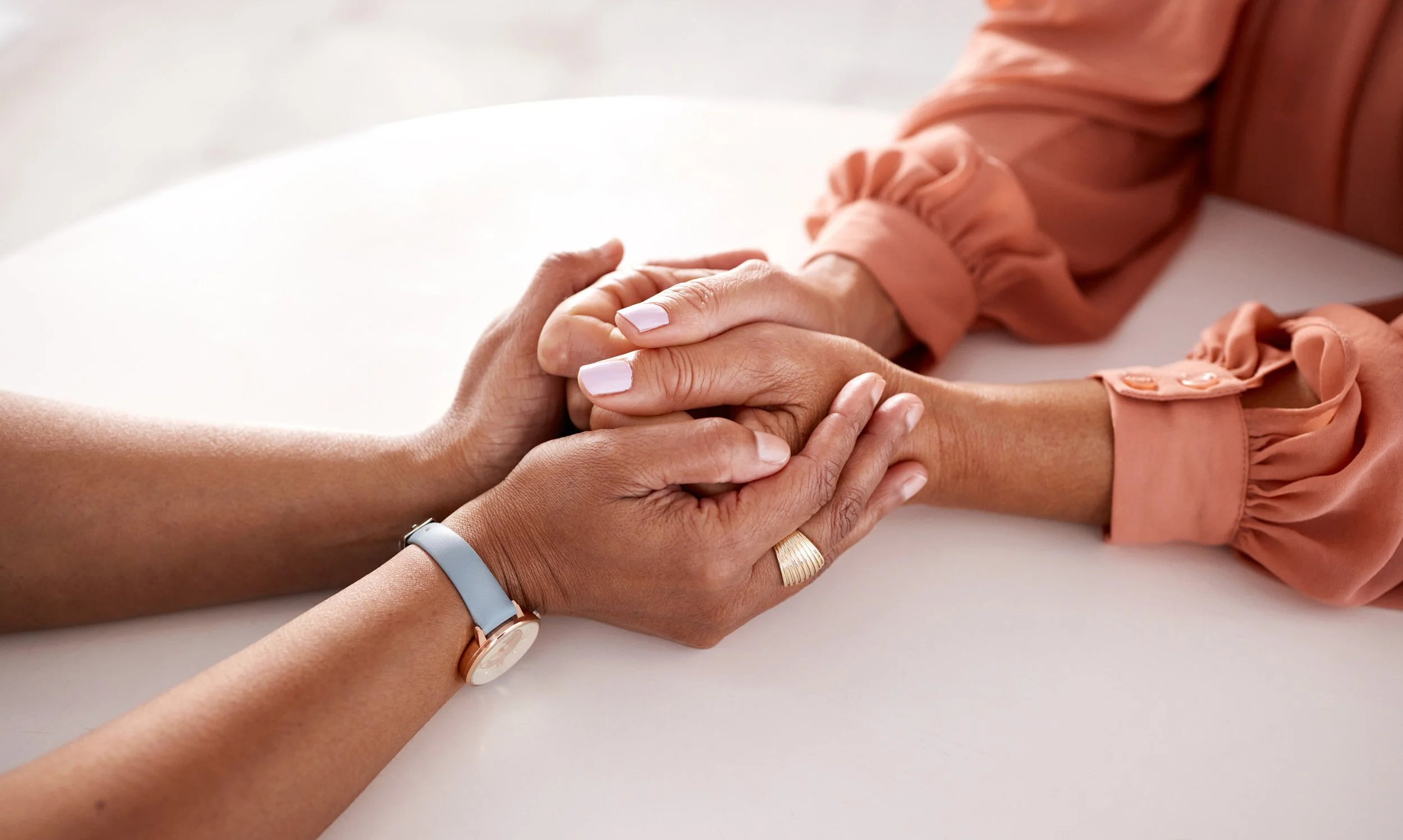 Two people holding hands on a white surface, one wearing a watch and a gold ring, the other in a pink blouse with puffed sleeves.