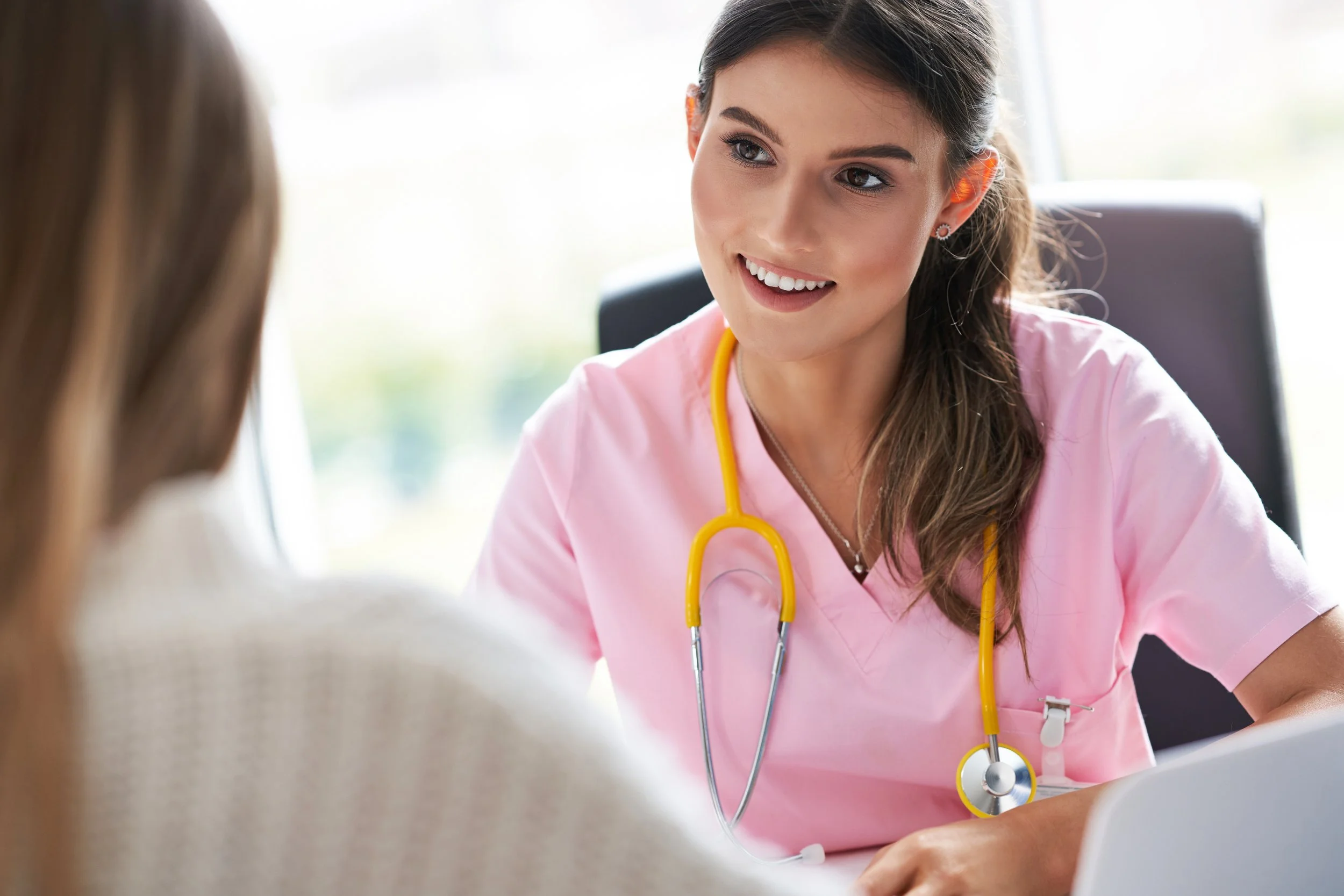 A young female nurse with a yellow stethoscope talking to a patient in a clinical setting.