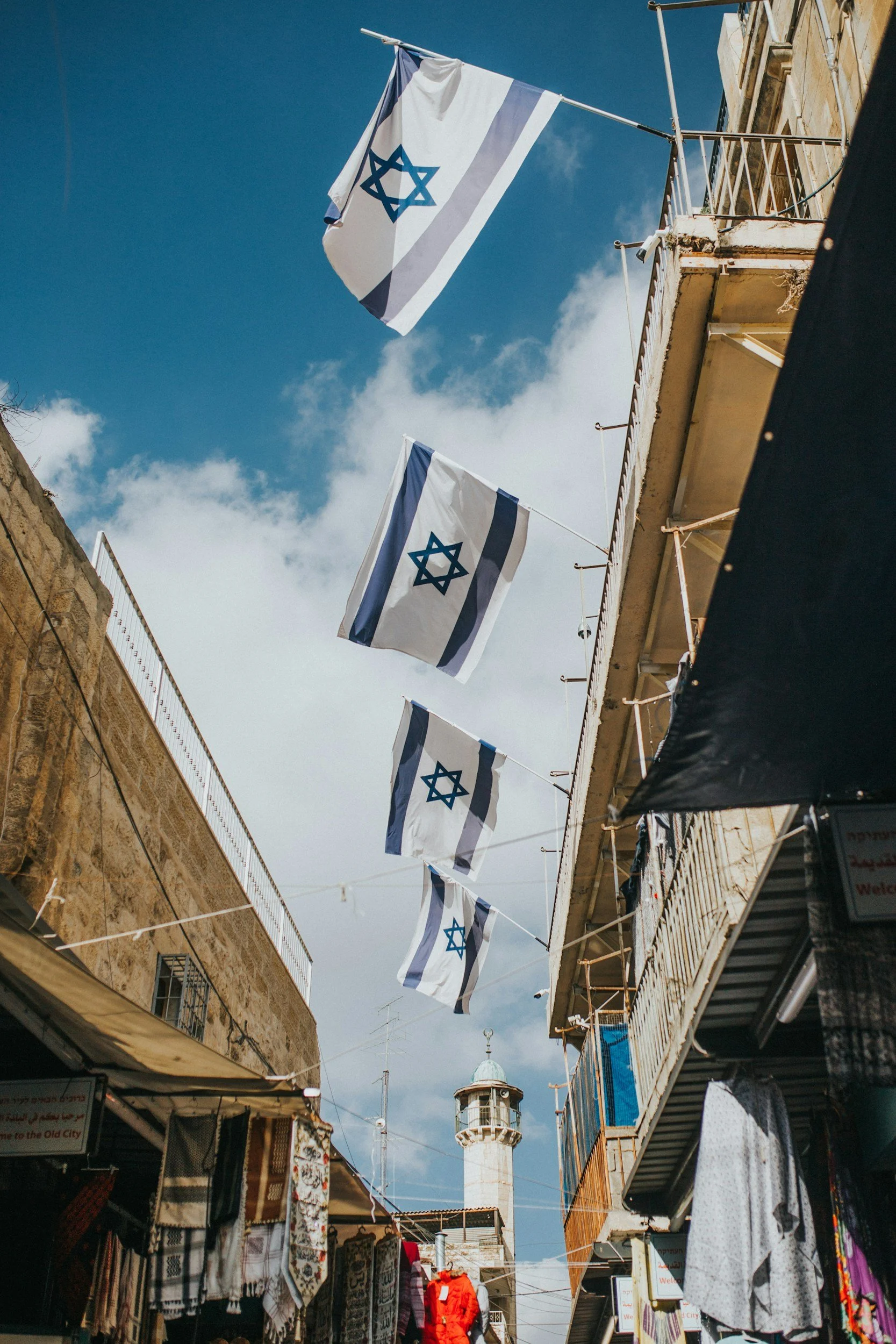 Three Israeli flags hanging between buildings in a market street with a minaret in the background.