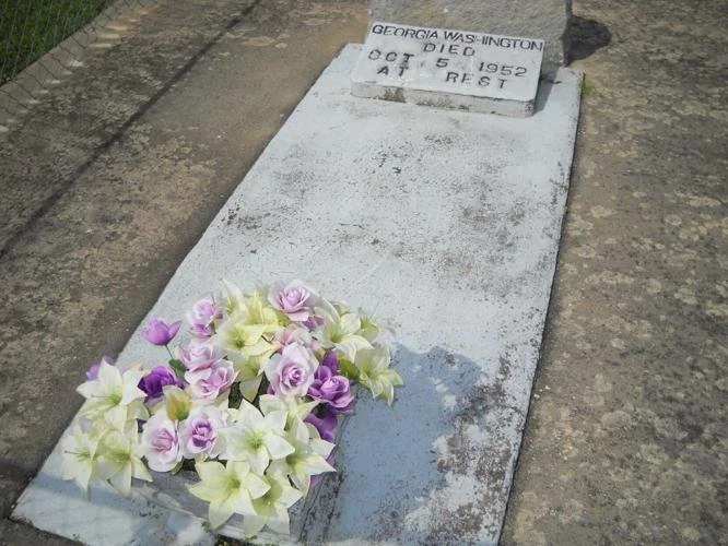 Grave marker for Georgia Washington with a white concrete headstone, decorated with purple and white flowers placed at the foot of the grave.
